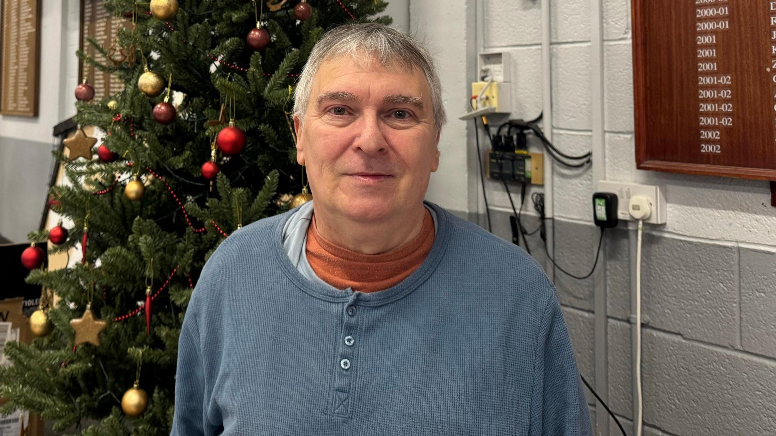 Gordon, a man with short hair, stands in front of Christmas tree wearing a blue button-up top