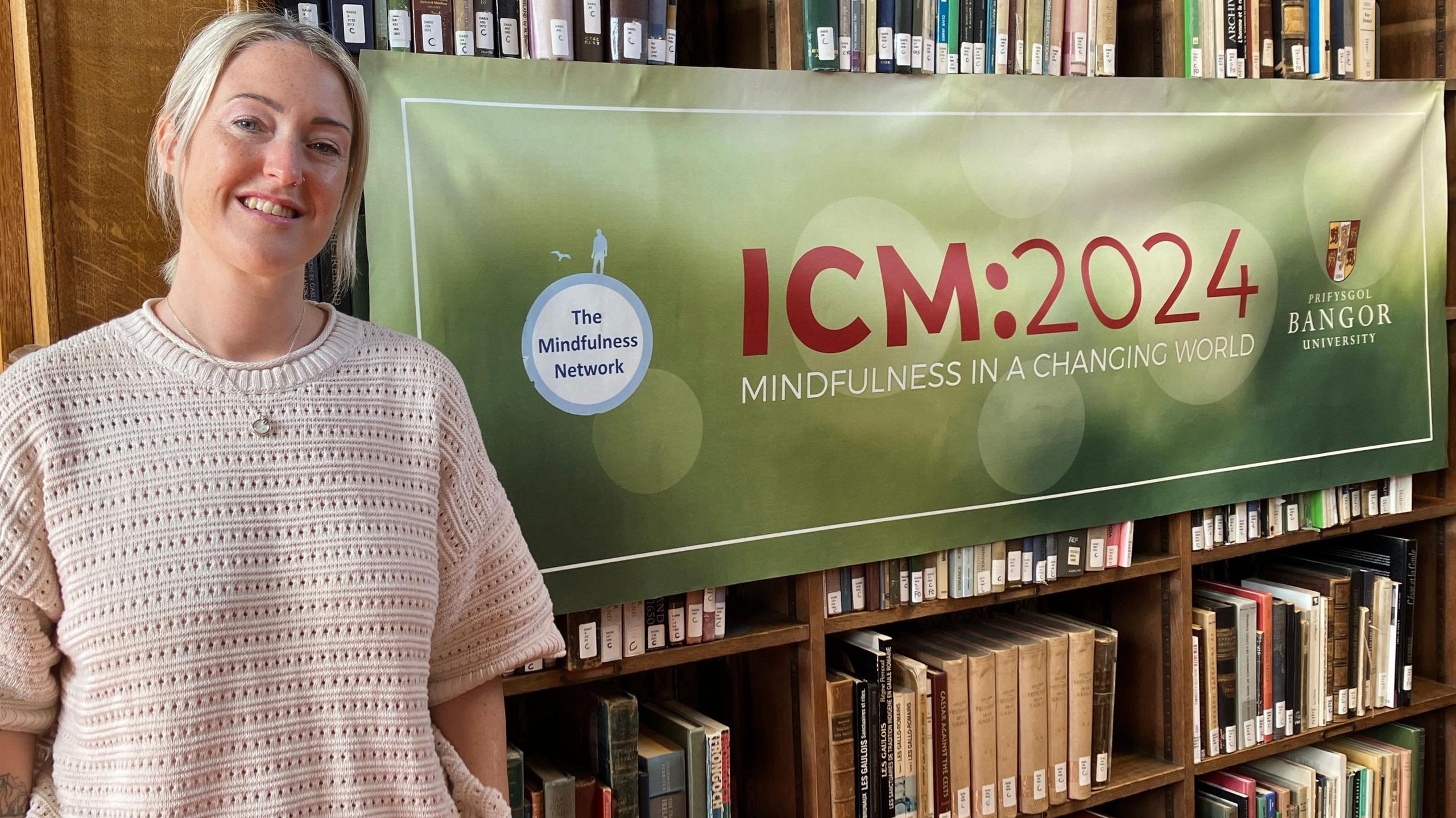 Esther Ghey, who has a cream jumper on, blonde hair and tattoos down one arm, standing in front of a bookcase, which has a green banner draped in front of it which says Mindfulness in a Changing World