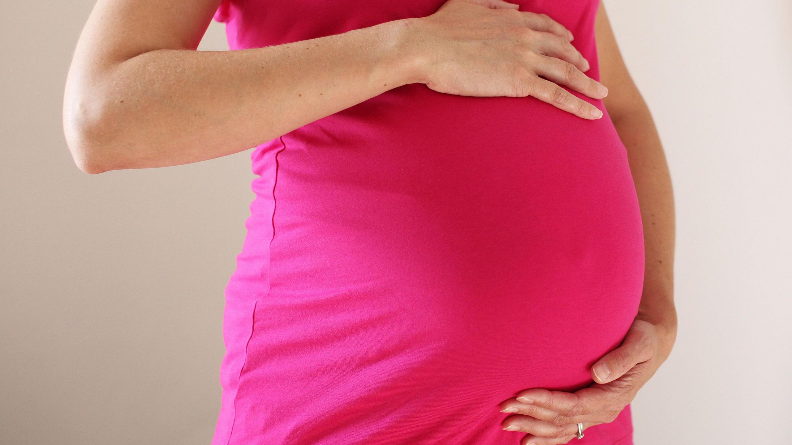 The torso of a woman wearing a bright pink dress who is eight months pregnant. She has her hands on the top and bottom of her baby bump.