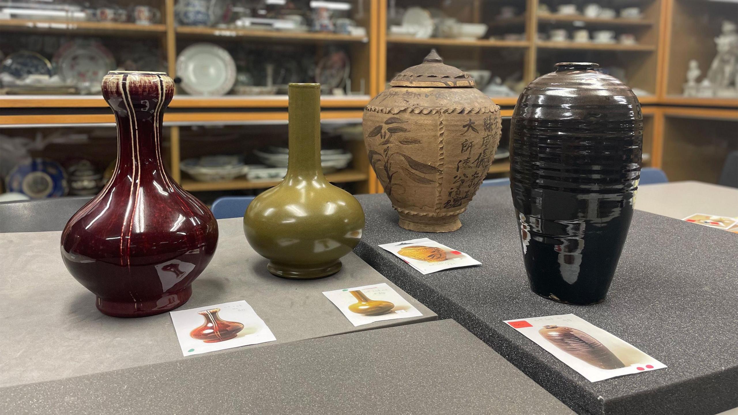 Four items from the Thornhill Collection of ceramics. They are various colours, and are positioned on a table with glass cabinets full of ceramics in the background.