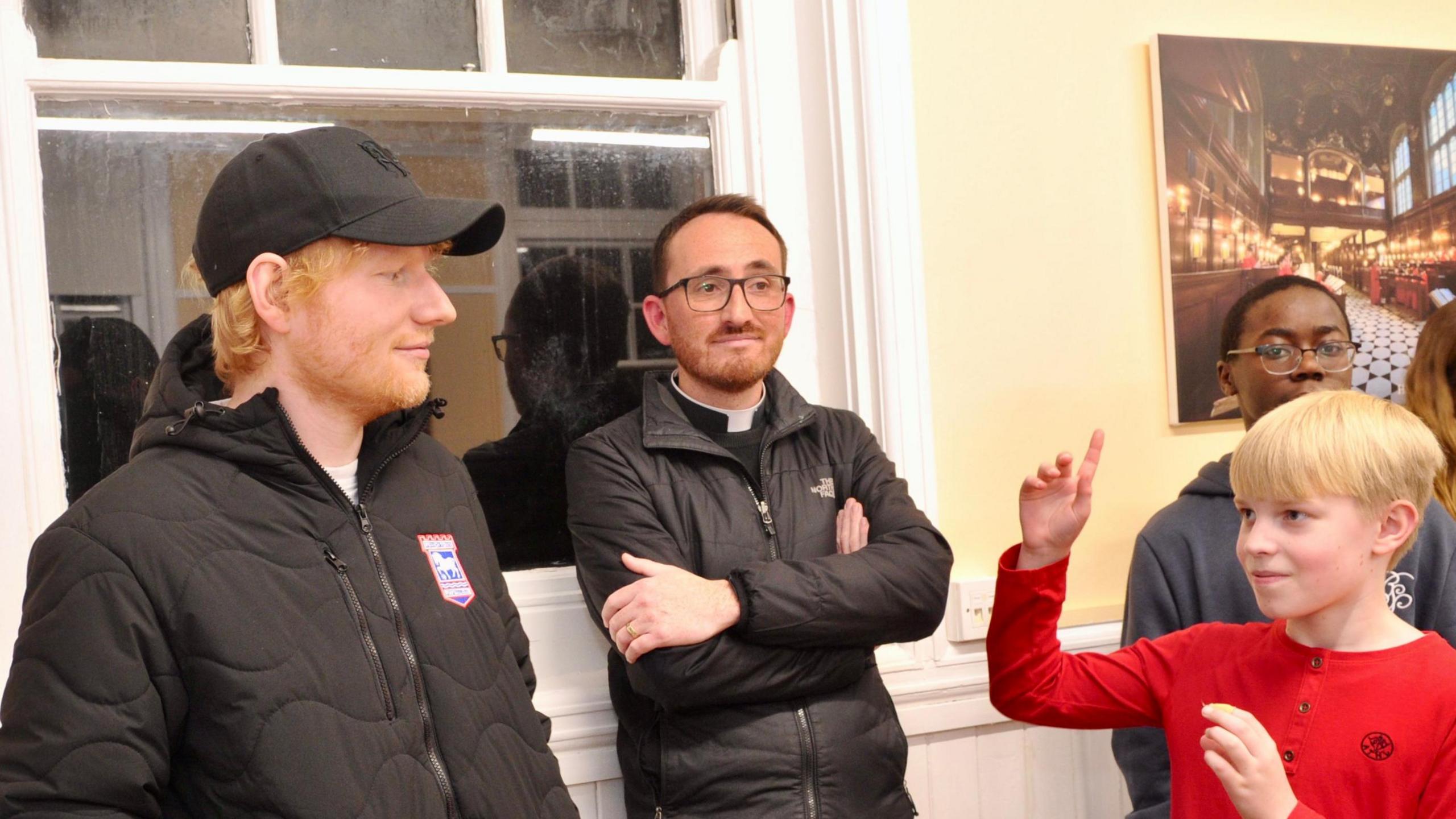 Ed Sheeran wearing a black cap and black Ipswich Town jacket and looking towards a fair haired boy, wearing a red top, and with his right hand raised. Between them leaning against a window is a vicar wearing his dog collar and leaning against a window frame with his arms crossed.