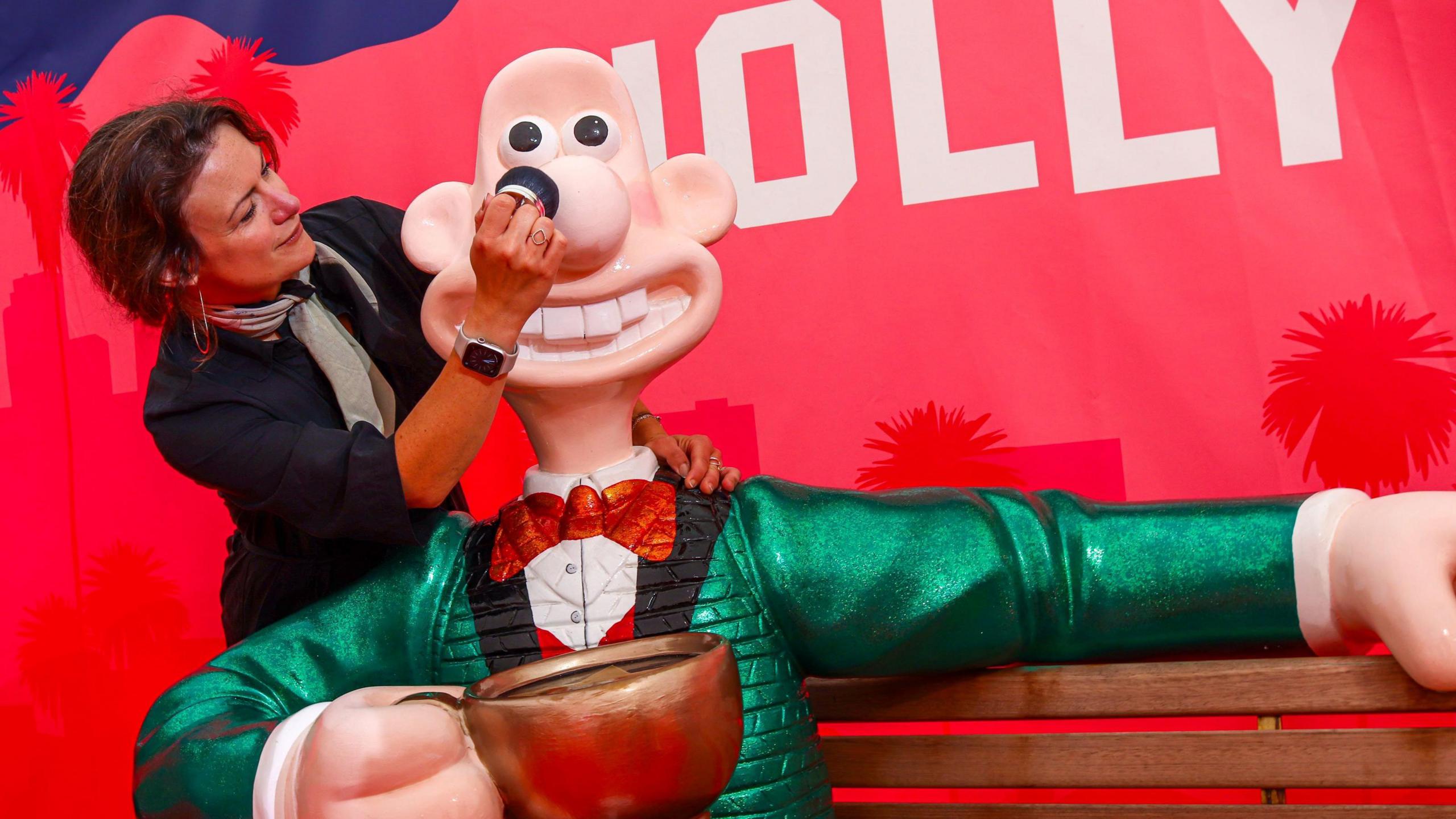 A young woman with brown hair and a grey scarf around her neck leans close to a sculpture of Aardman character Wallace, which is part of a display of Gromit Unleashed 3 at Cribbs Causeway in Bristol. The Wallace sculpture is wearing a green jacket and red bow tie and is holding a large cup of tea.