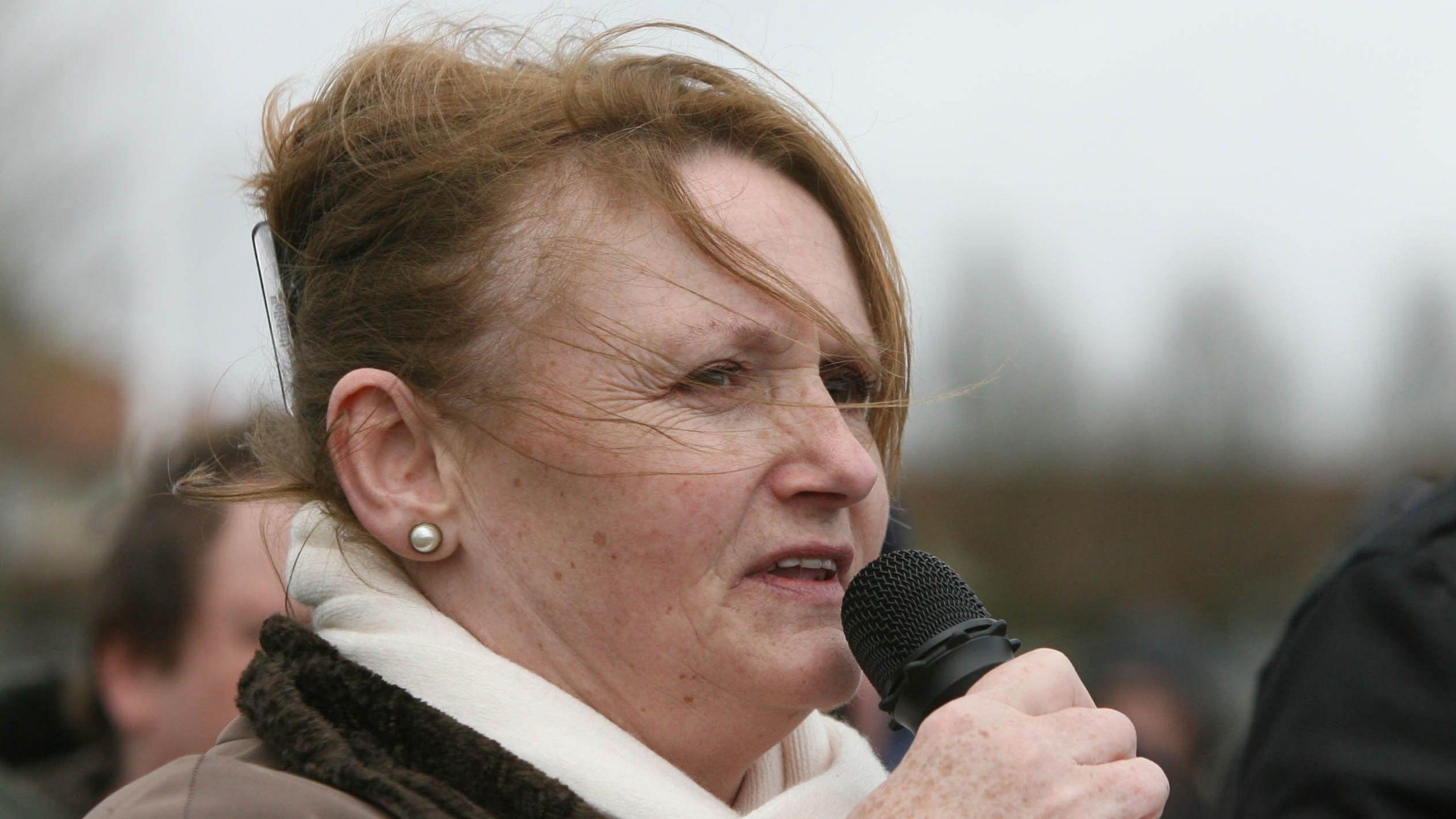 Marian Price holding a microphone, and wearing a white scarf. Her coat is brown and she is also wearing pearl earrings