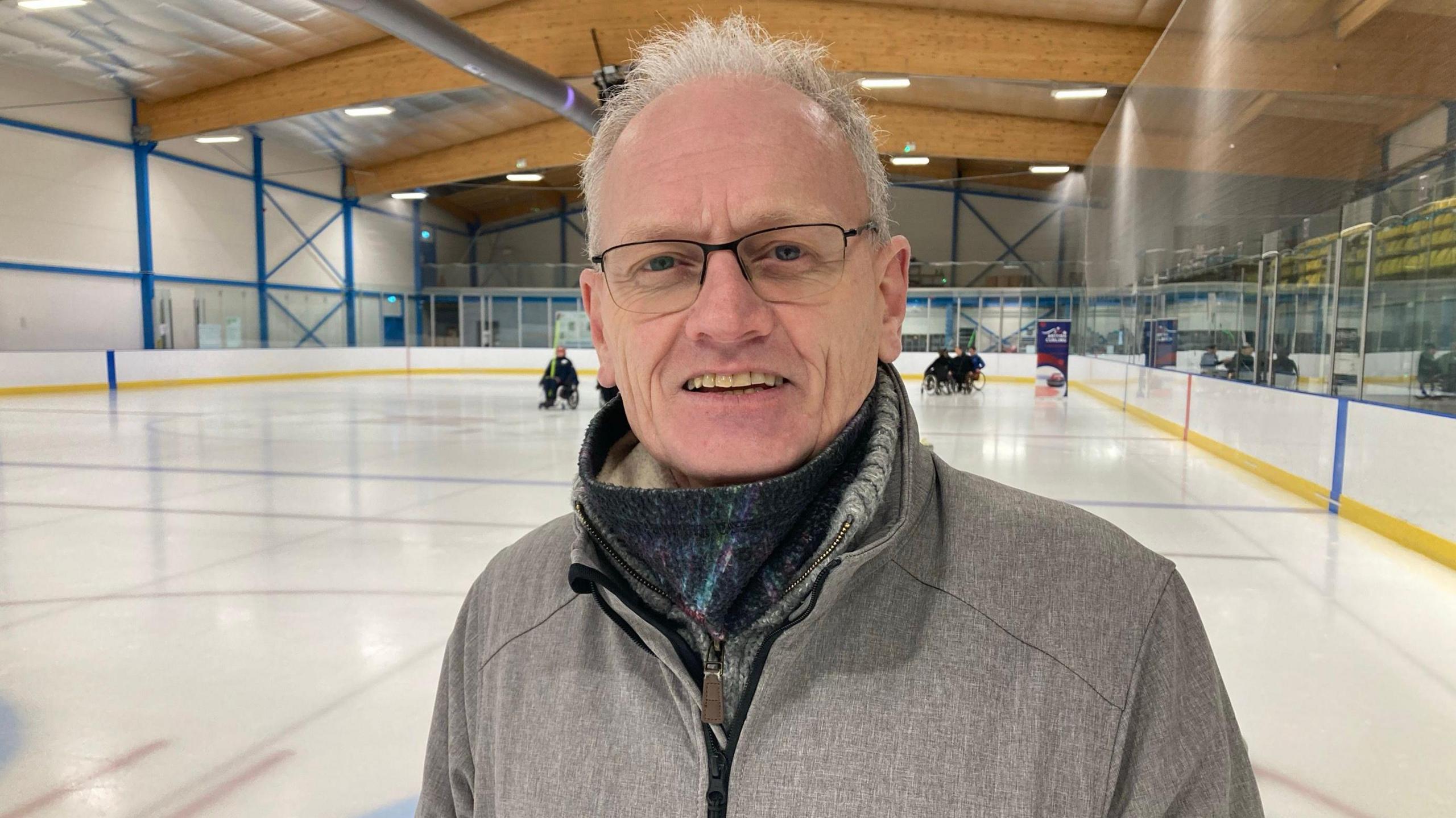 Stan Wilson, president of Cambridge Curling Club is standing on the ice rink looking at the camera. He is wearing a grey top and has grey hair and glasses. People can be seen behind him on the rink in wheelchairs