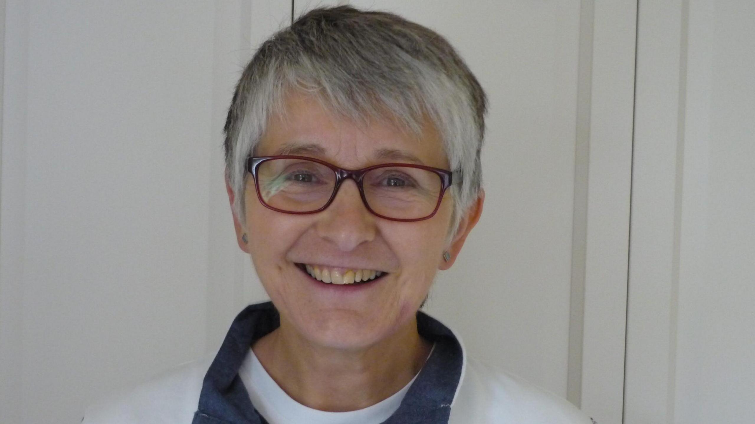 Sarah Lovegrove smiles at the camera, wearing glasses and a white jumper with a blue collar