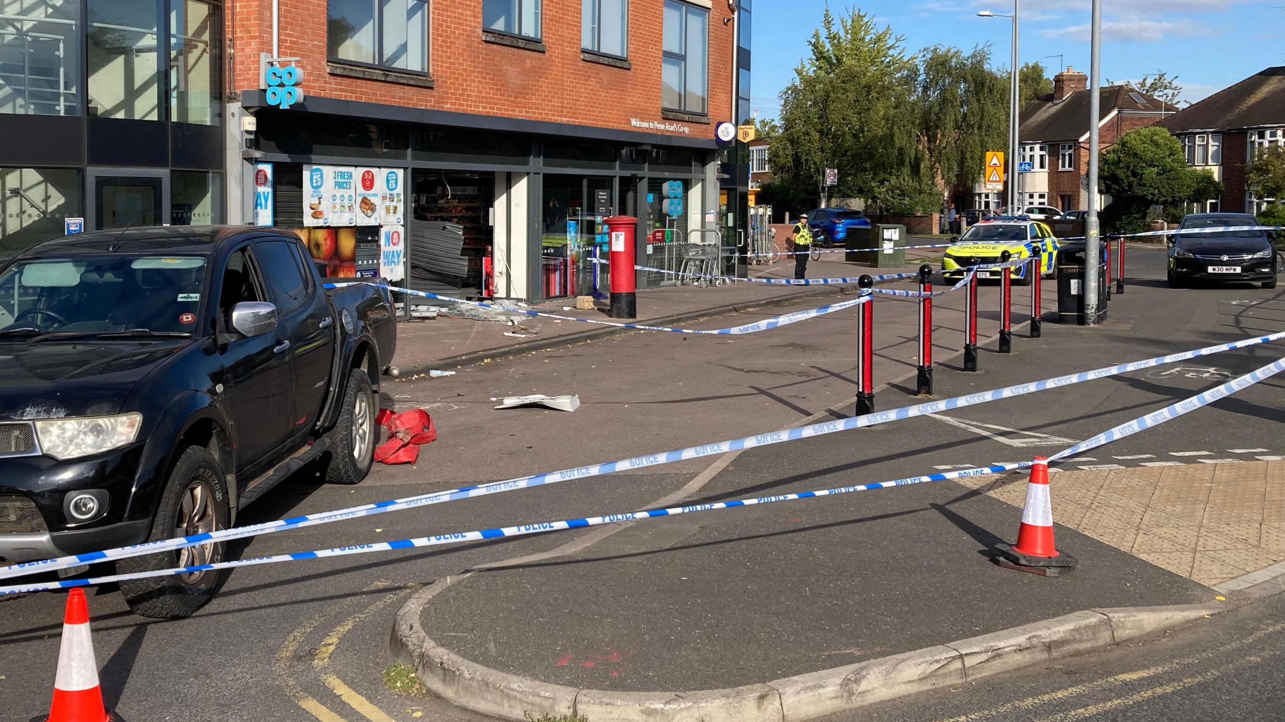 A large police cordon has sectioned off the front of the coop and includes the black car - believed to have been involved in the ram raid. At the back of the car are thick red straps hanging off the back. The front of the shop has been smashed and there is glass on the floor. The shutters have been hit and pushed inwards, leaving the entrance to the hop wide open.