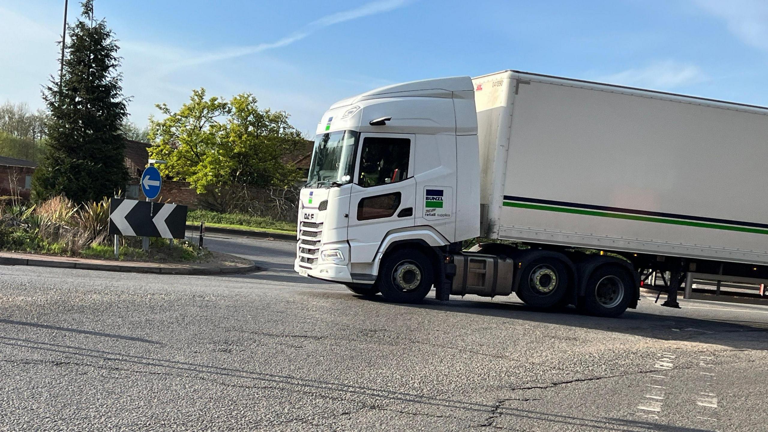 A heavy goods vehicle with a white cab and trailer approaching a roundabout. There are some clouds in an otherwise clear, blue sky.
