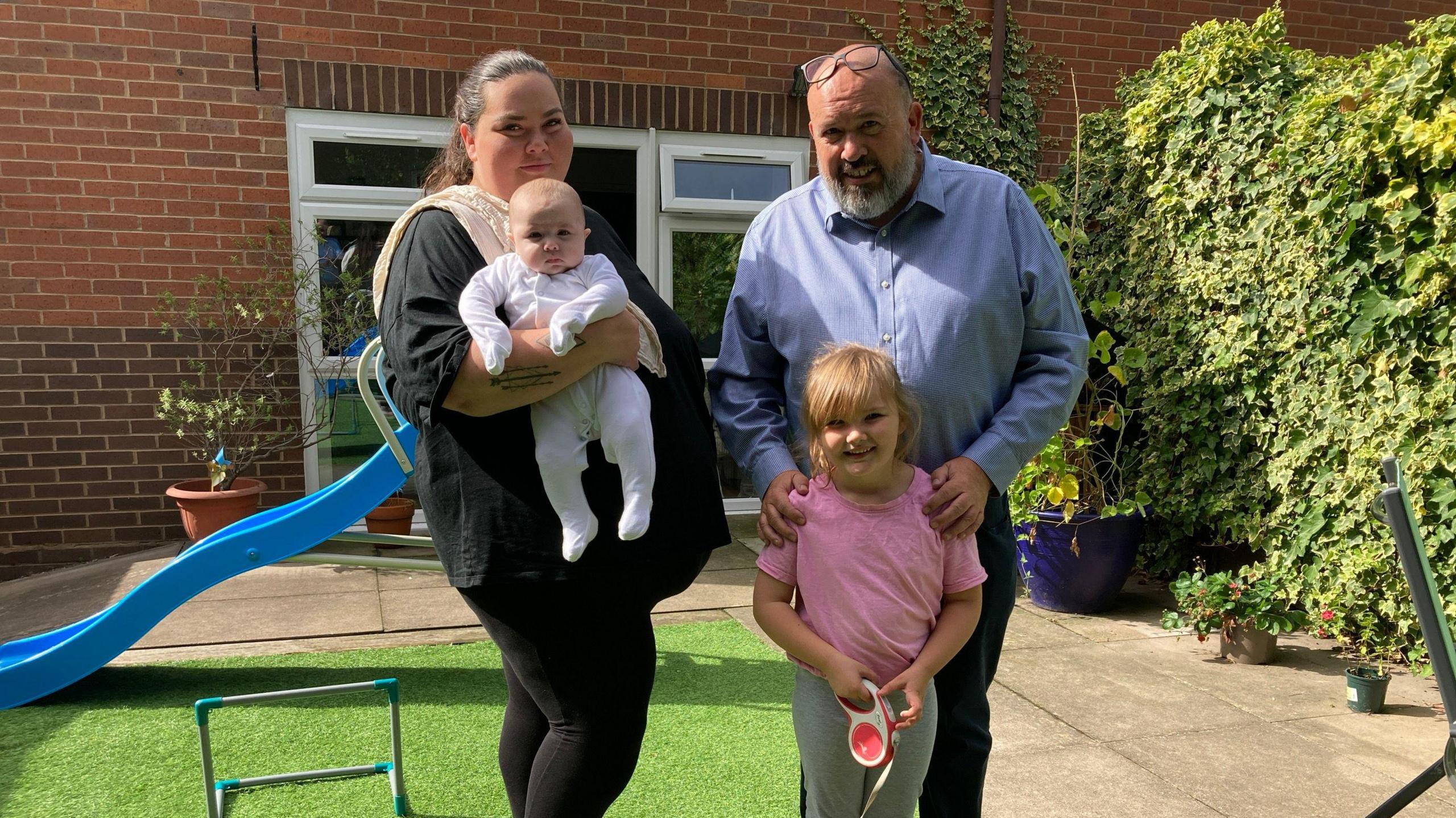 A woman in a black top and leggings holds a baby, while standing next to a man with a grey beard and a young girl wearing a pink t-shirt. The family are standing in a back garden.