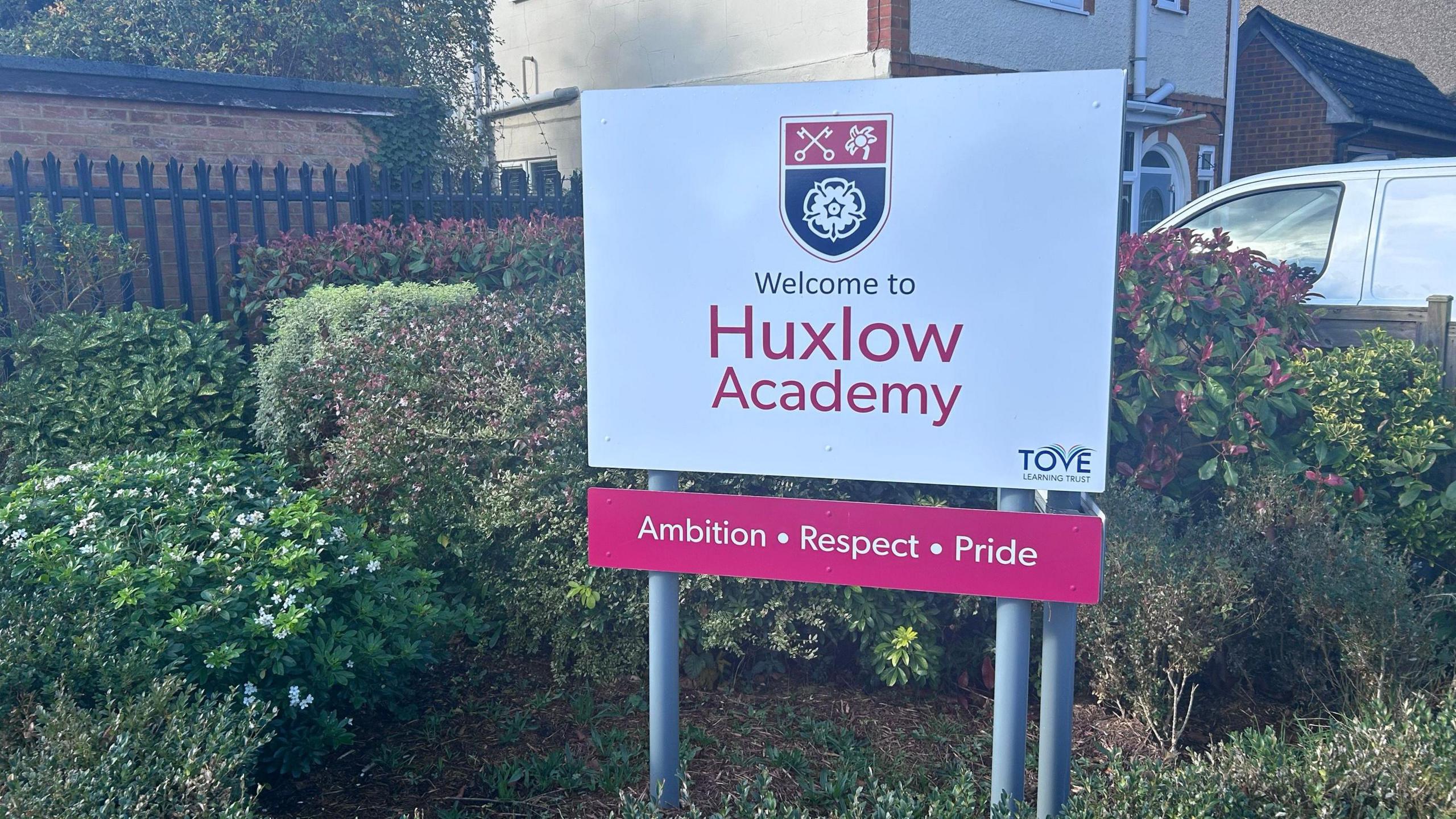 A white free-standing sign outside the school says "Welcome to Huxlow Academy" and shows the school crest, which has a set of crossed keys and two flowers. Below is says Ambition; Respect; Pride. The sign is next to a residential house with a van parked outside.