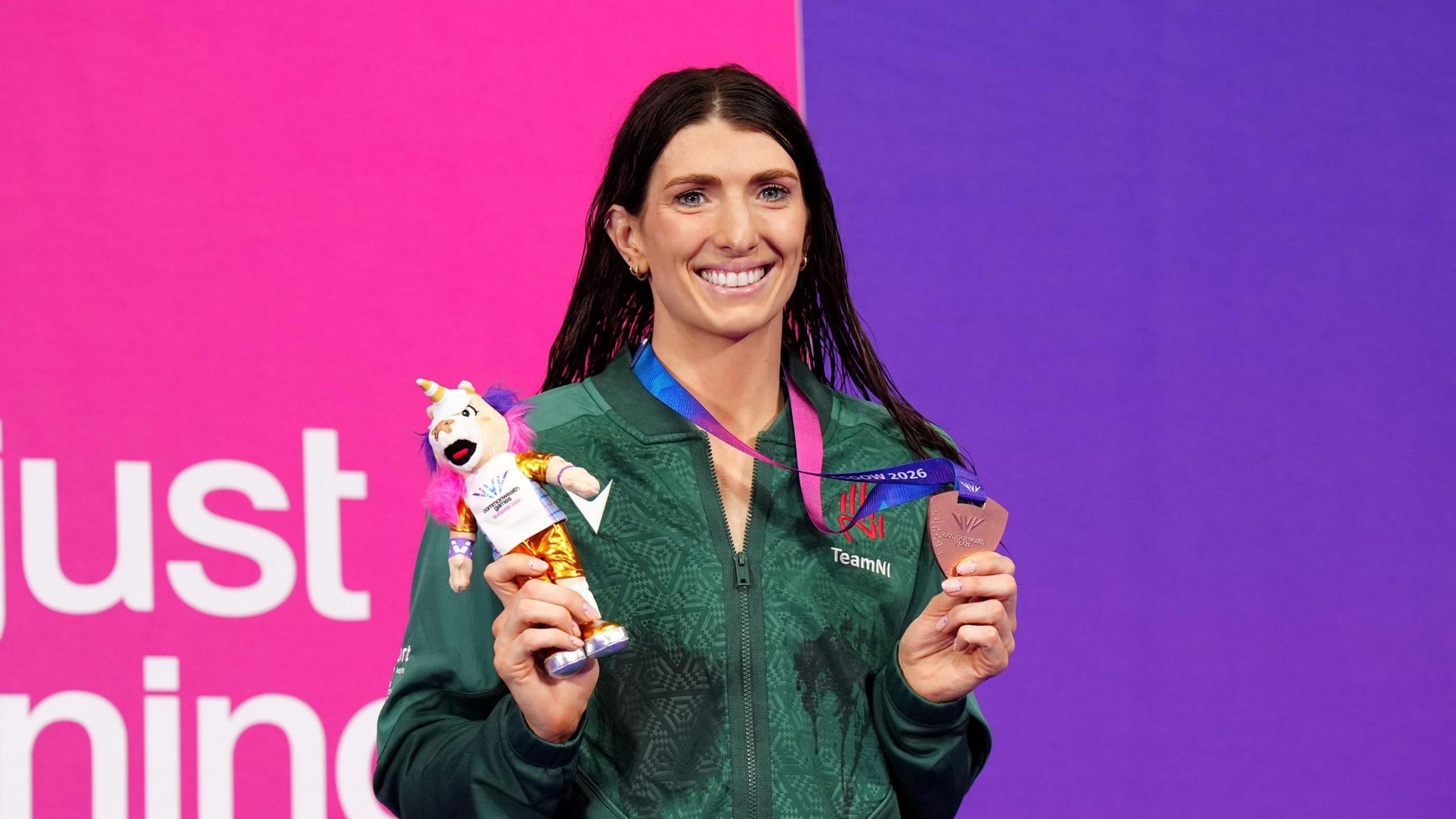 Bethany smiling holding her medal and a small commonwealth games mascot. She is wearing a green team NI jacket. She has long brown hair.