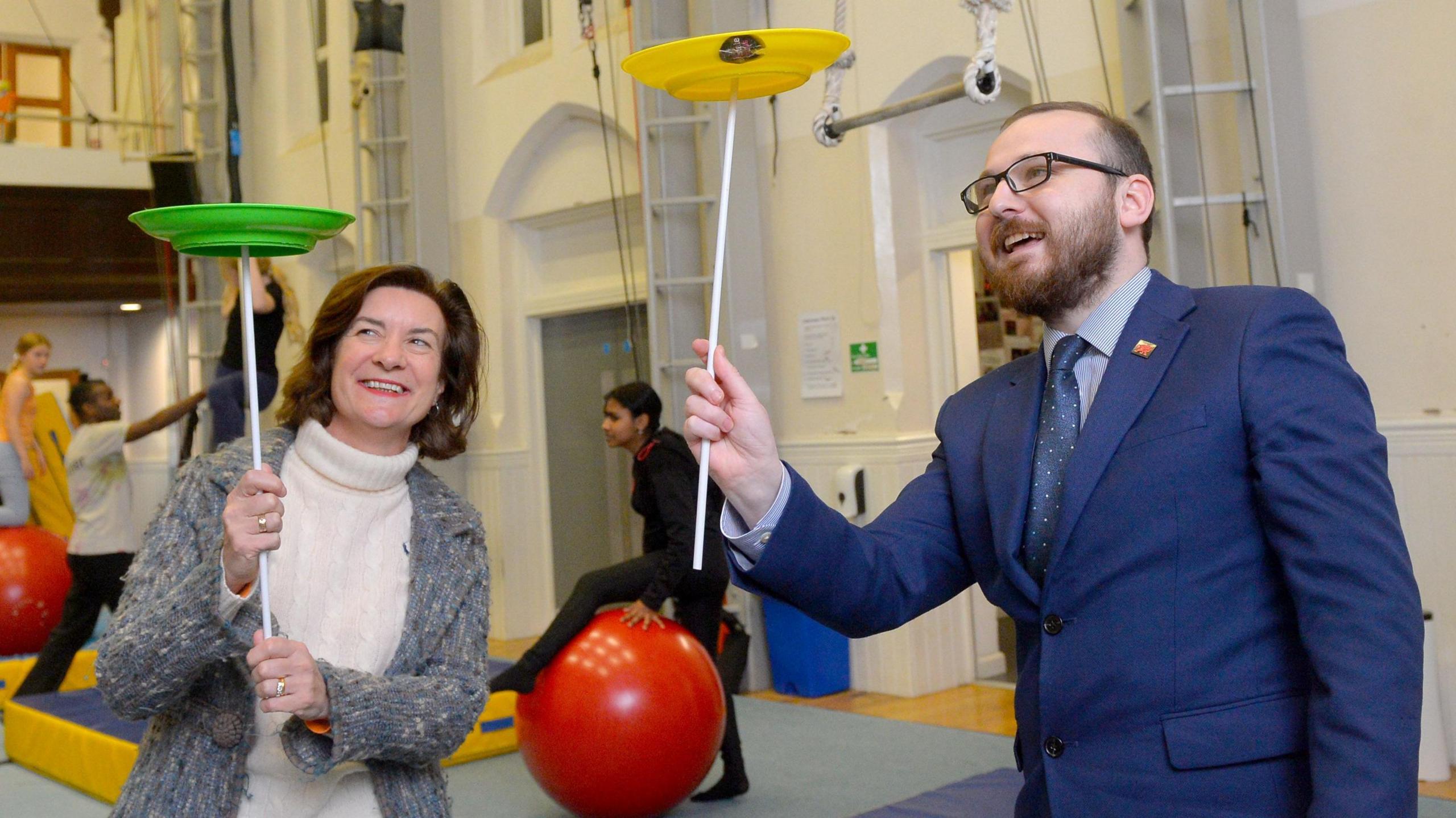 First Minister Eluned Morgan and Culture Minister Jack Sargeant spinning plates during a circus workshop. Eluned Morgan is holding a green plate on a stick while Jack Sargeant is holding a yellow plate.