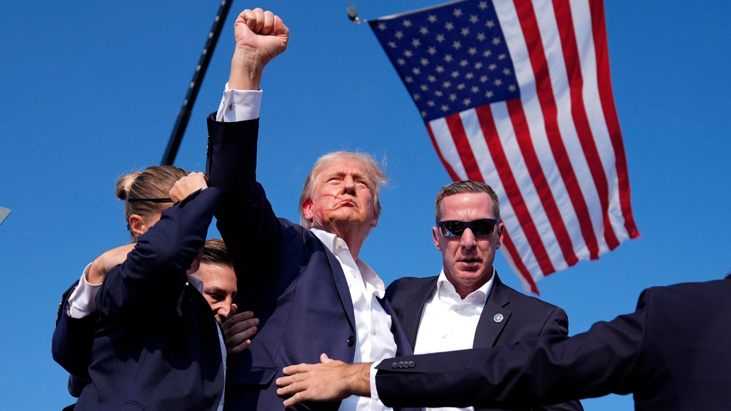 Trump, with blood smeared across his face, raises his fist as he leaves a campaign stage after being shot at. He is surrounded by US secret service agents. A US flag is flying in the background.