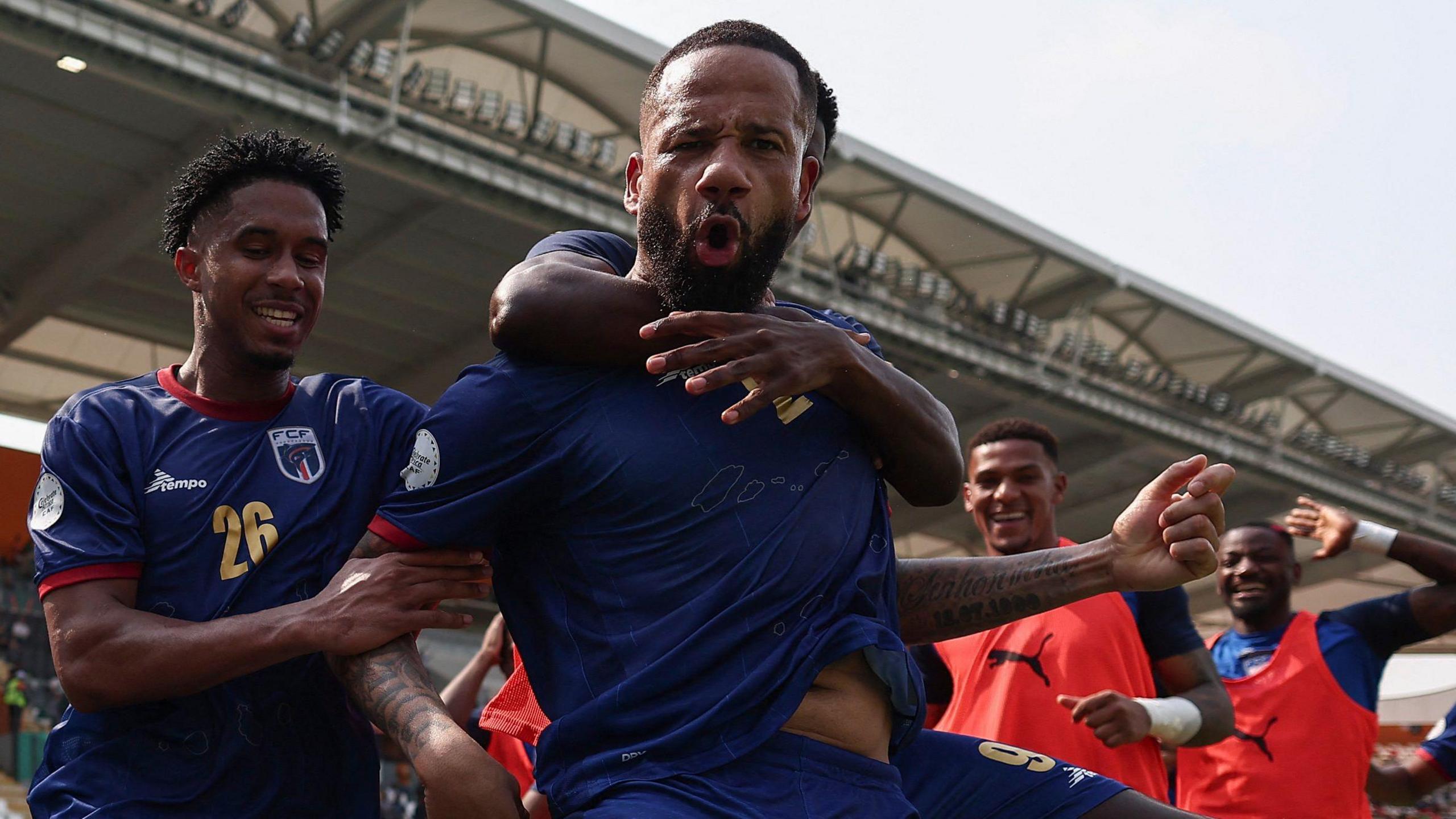 A bearded Bebe pulls a face as he looks down into the camera positioned at a low level while celebrating a goal at the 2023 Africa Cup of Nations. He is wearing dark blue kit which is being pulled by a couple of team-mates who are grabbing him from behind, with one putting both arms around Bebe's neck. Two more smiling team-mates wearing orange substitute bids run towards the group while the roof of a stand is in the background