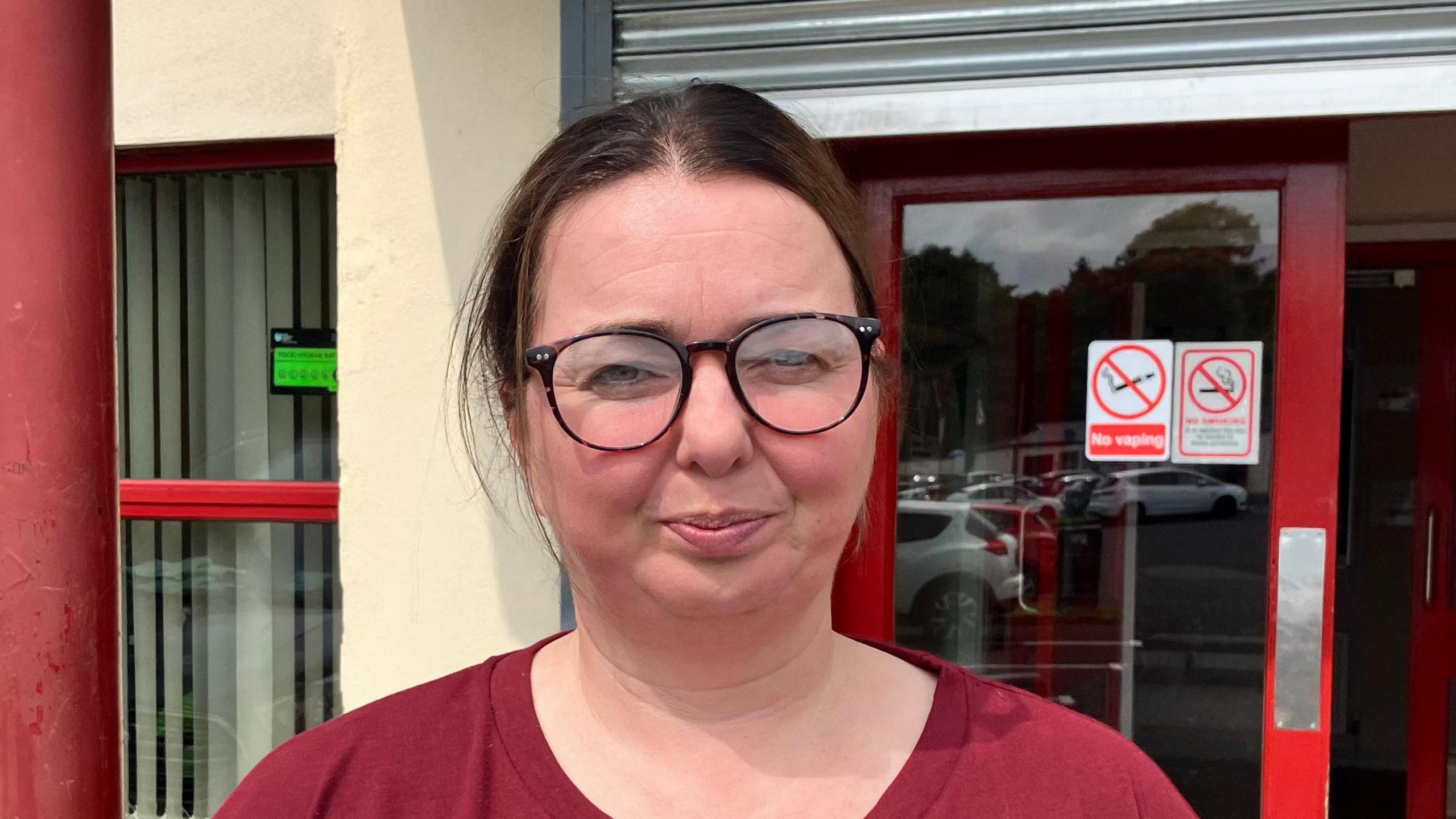 A lady with brown hair tied back. She is wearing black round-lens glasses. Behind her is red doors and a cream building.