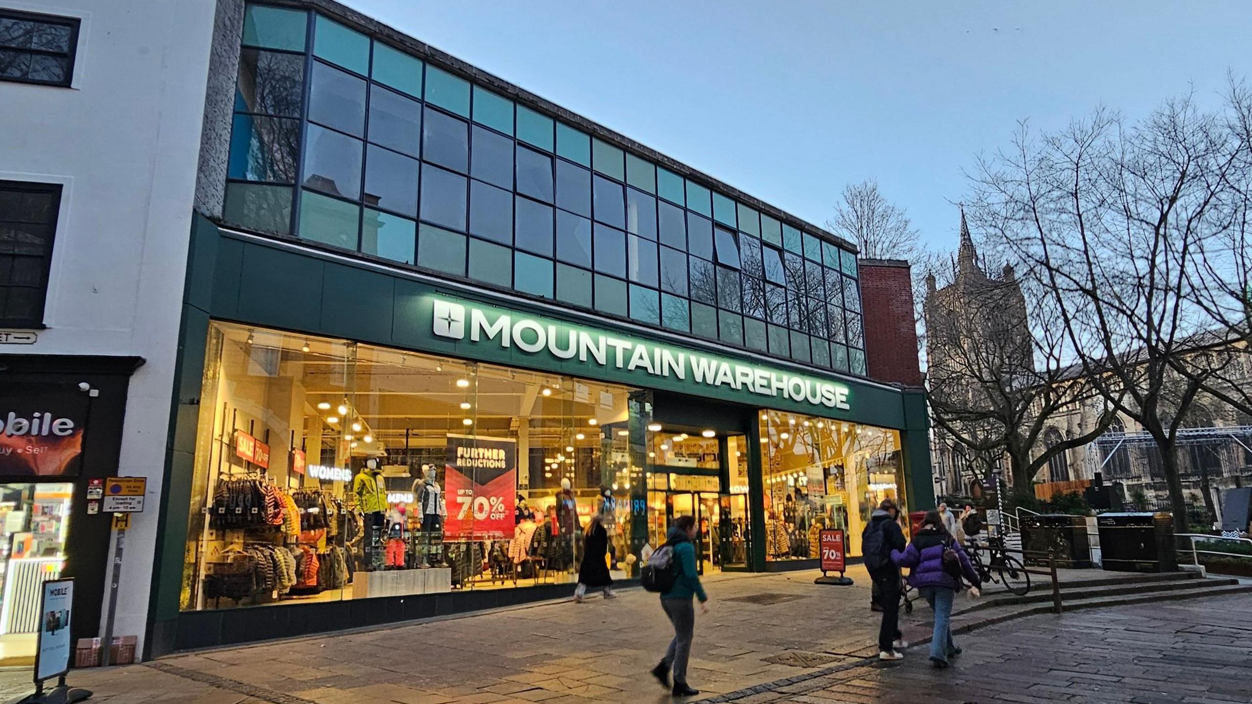 A picture of the front of Mountain Warehouse. Daylight is fading so it the shop is lit up, with a number of pedestrians walking past.