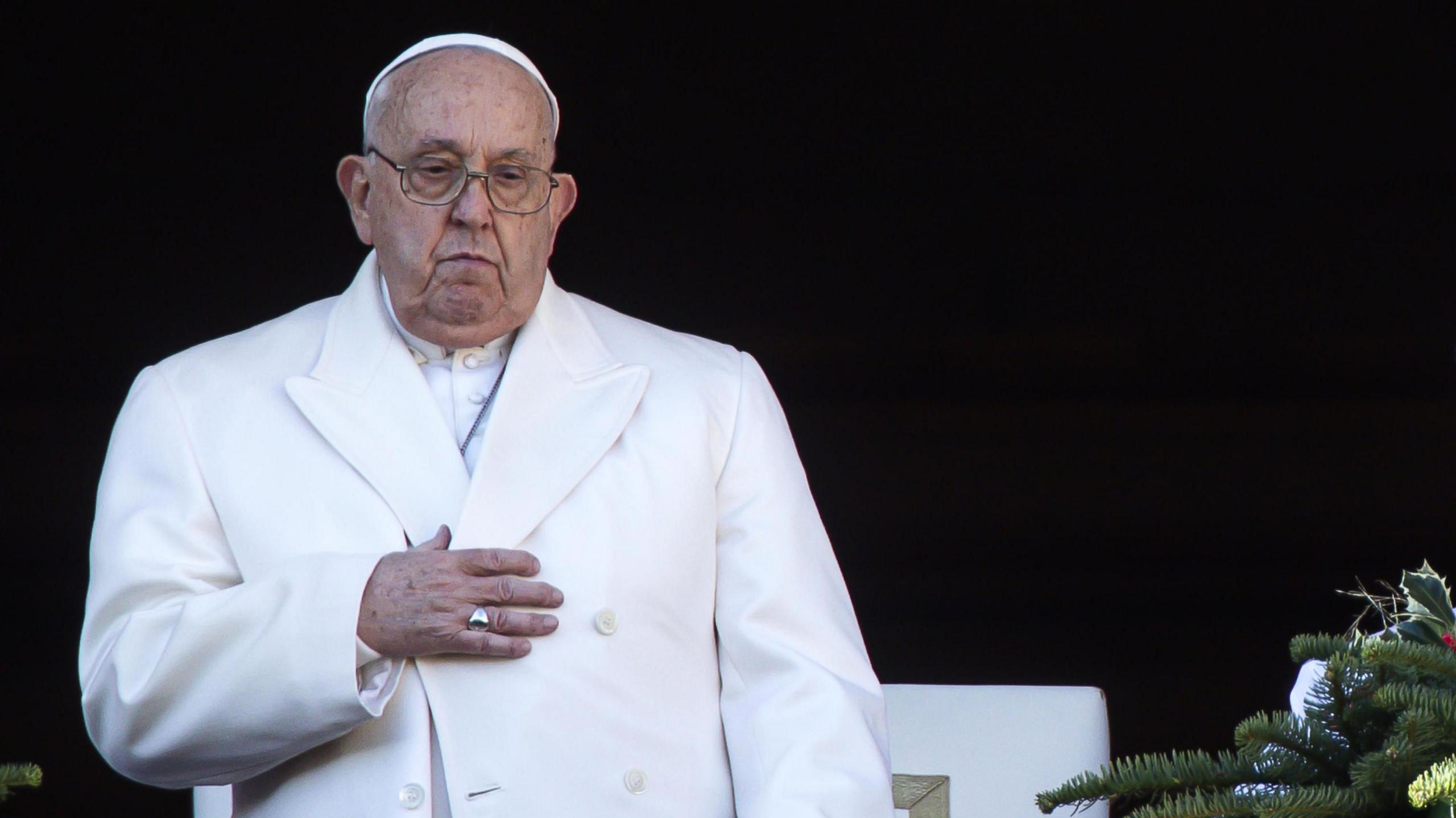 Wearing a white coat the Pope touches his chest while standing up and looking down at a crowd on Christmas Day