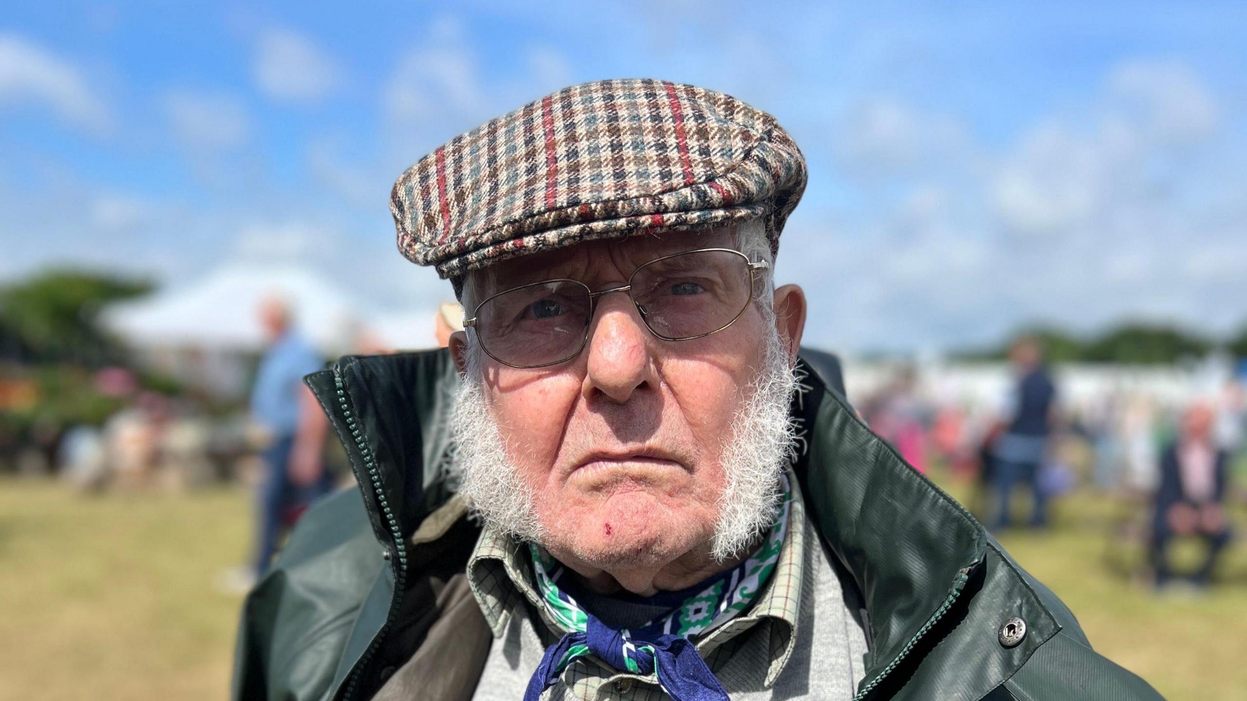 A close-up photo of an elderly man wearing a flat cap and a neck scarf. He is looking directly into the camera.