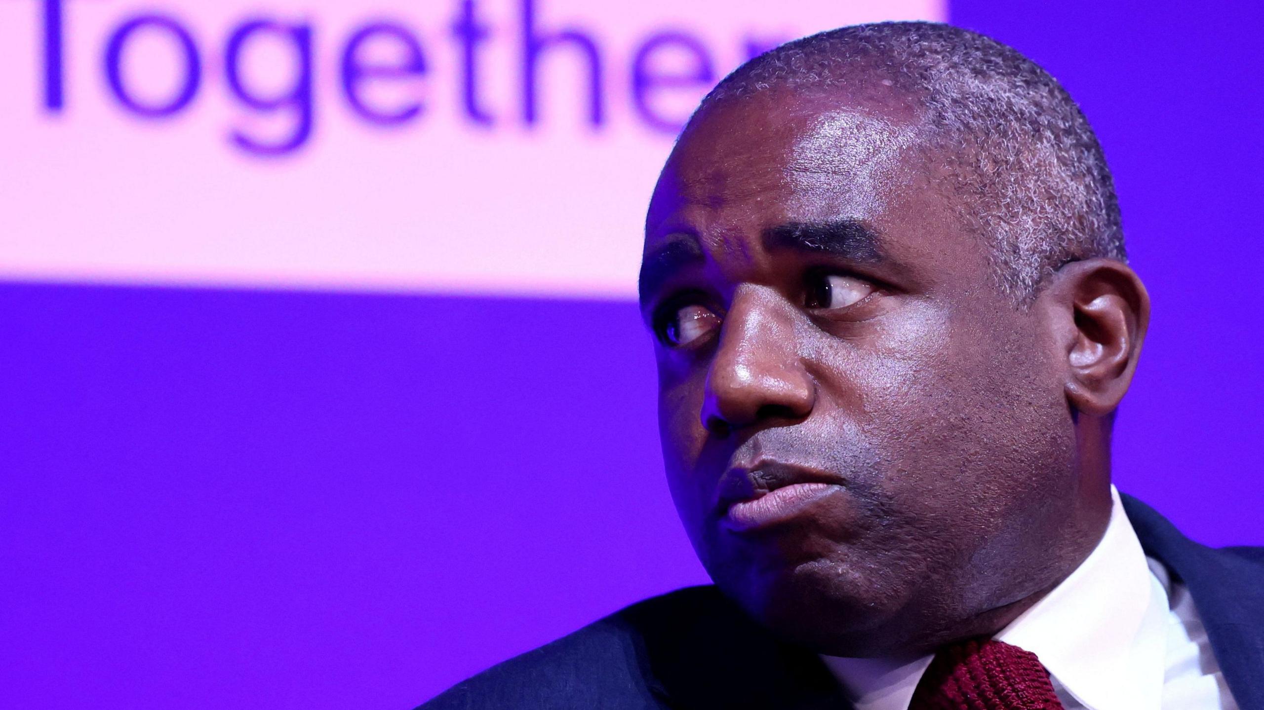 David Lammy, wearing a suit and maroon tie, with a purple background saying 'together' - he is looking to his side