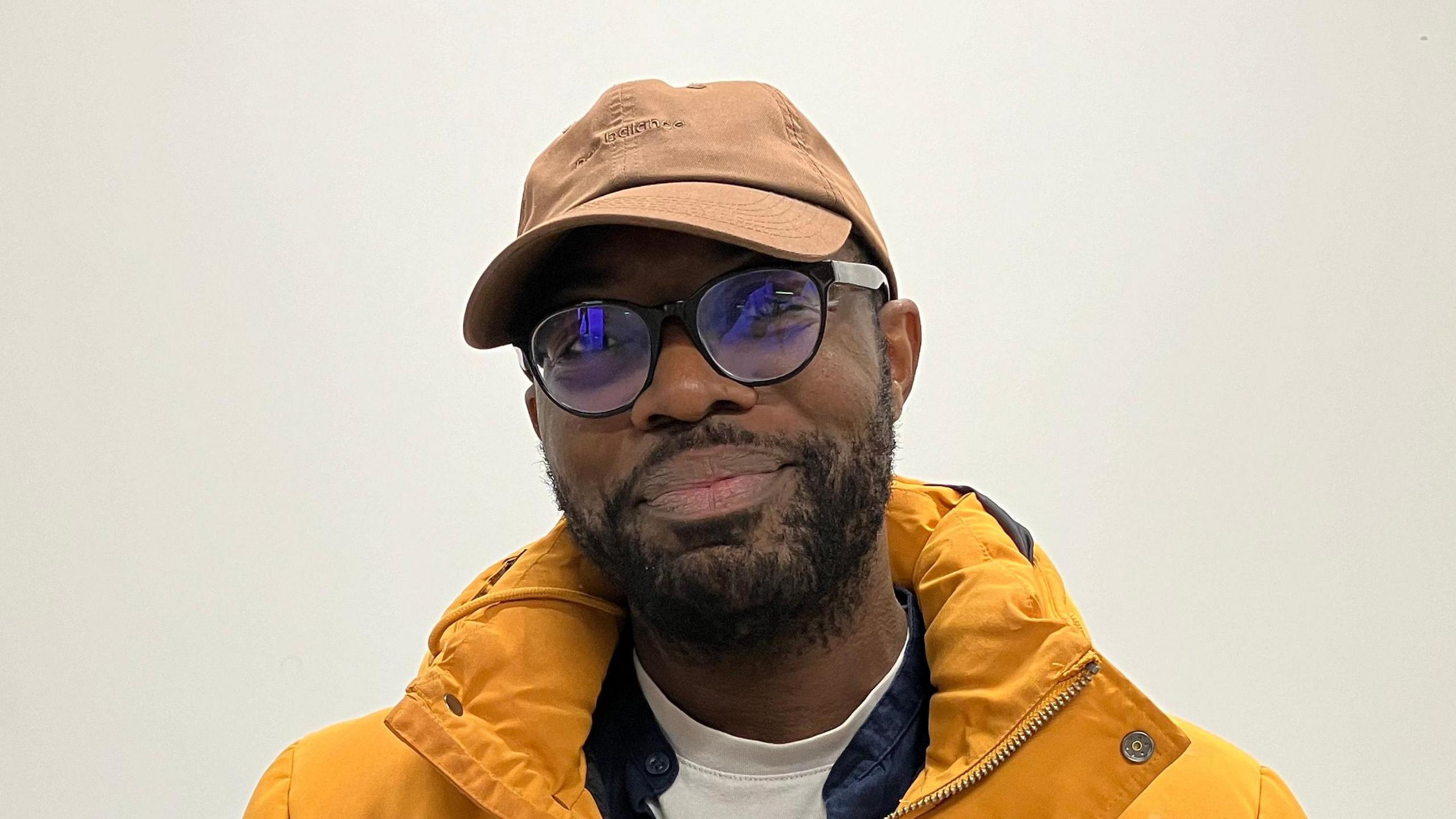 Nana Parry smiles at the camera. He wears a brown cap with black glasses and a yellow puffer coat. 