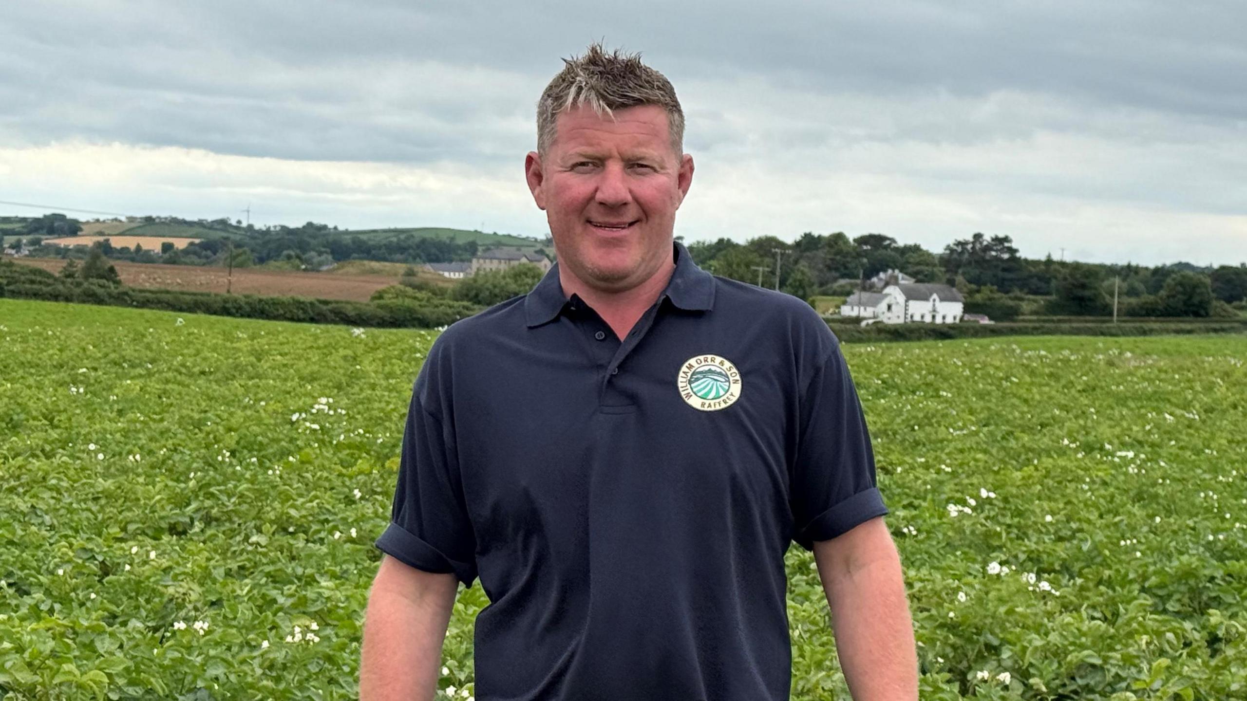 Richard Orr standing in front of potato crops. He has short brown hair and is wearing a navy polo shirt. There are hills and a white-coloured house in the distance behind him.