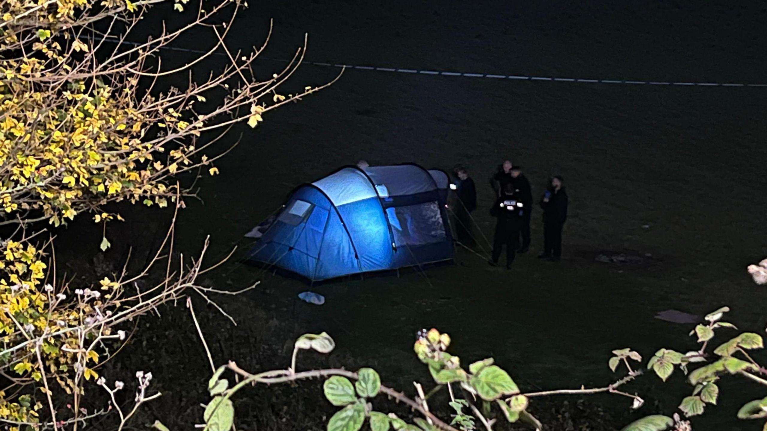 Six uniformed police officers on a field next to a tent, which is lit inside