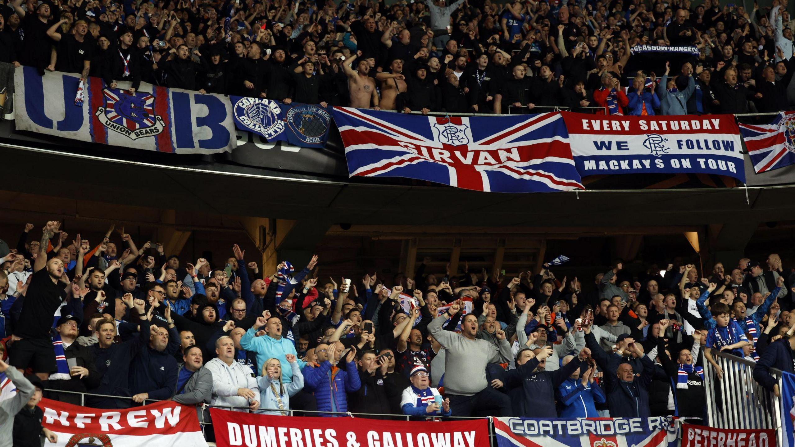 Rangers fans at the match with Nice, many dressed in replica kits. Many of the supporters are chanting and singing, while banners are draped around their section listing which area they are from. 