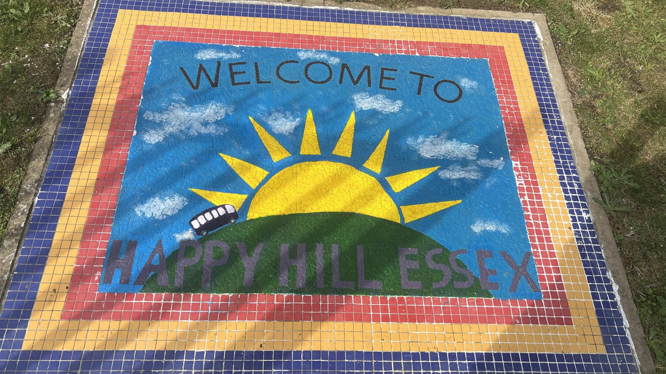 A colourful mosaic in the grounds of the Happy Hill charity's centre. In a blue, yellow and red border it shows the sun rising over a green hill. It reads: "Welcome to Happy Hill Essex"