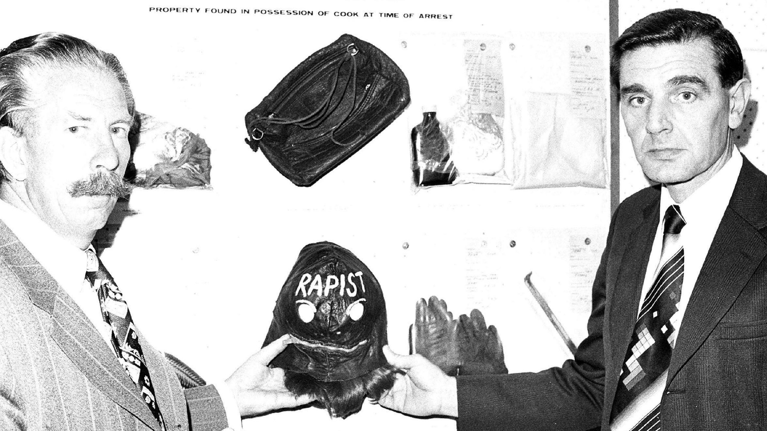 A black and white photo of two police detectives in suits holding a black leather mask with the word "rapist on it". Behind them are other items of evidence, including gloves, a handbag and medicine bottle.