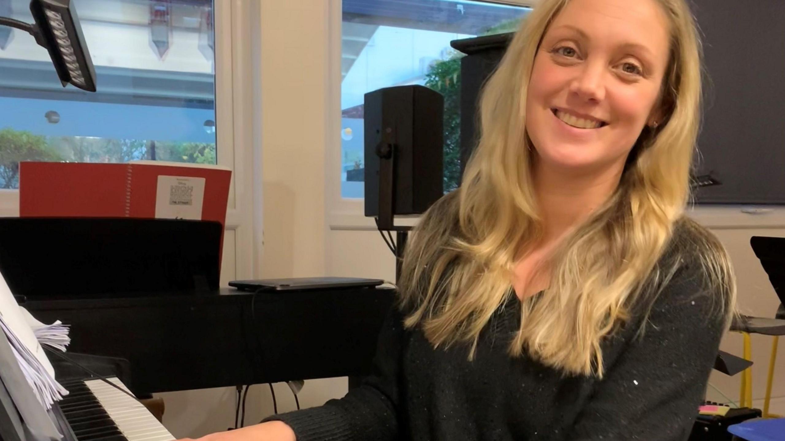 A smiling blonde woman wearing a black jumper is sitting down and playing a piano.