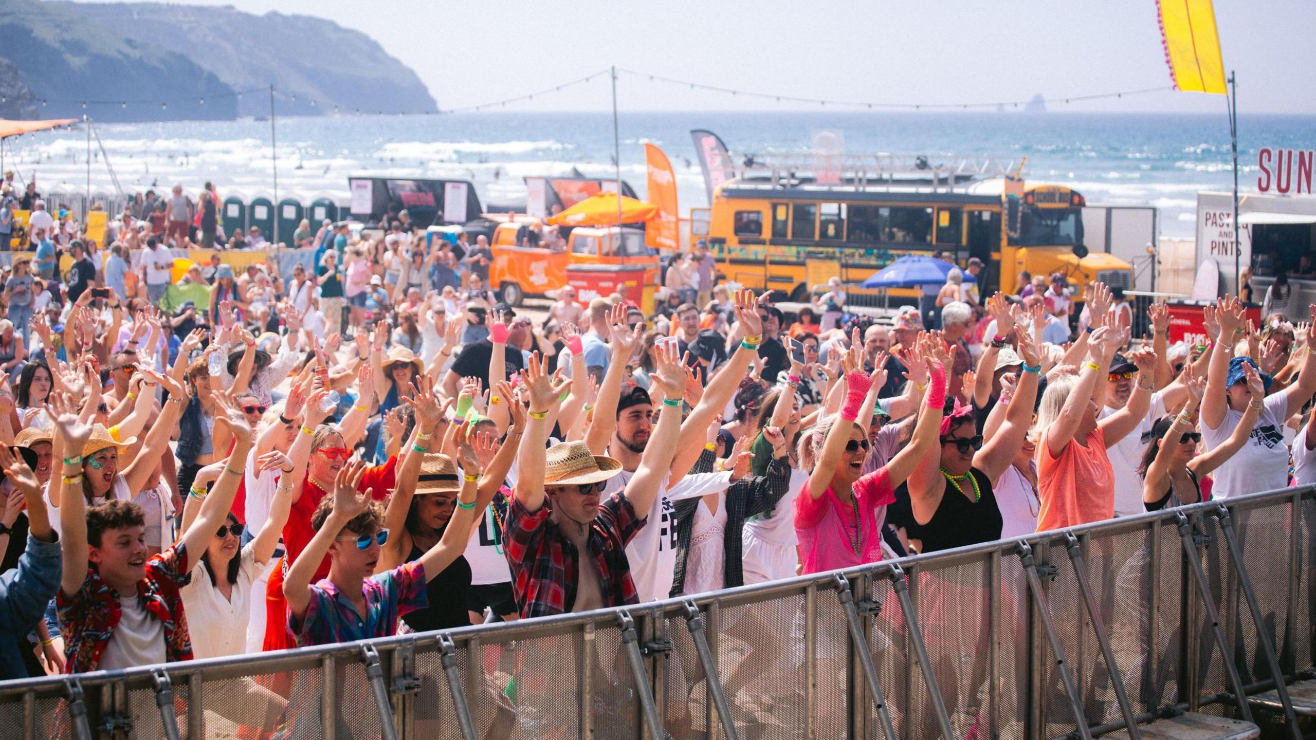 Hundreds of people stand at the barrier of a music festival on a beach with their hands in the air. It is a sunny day and you can see the sea in the background.
