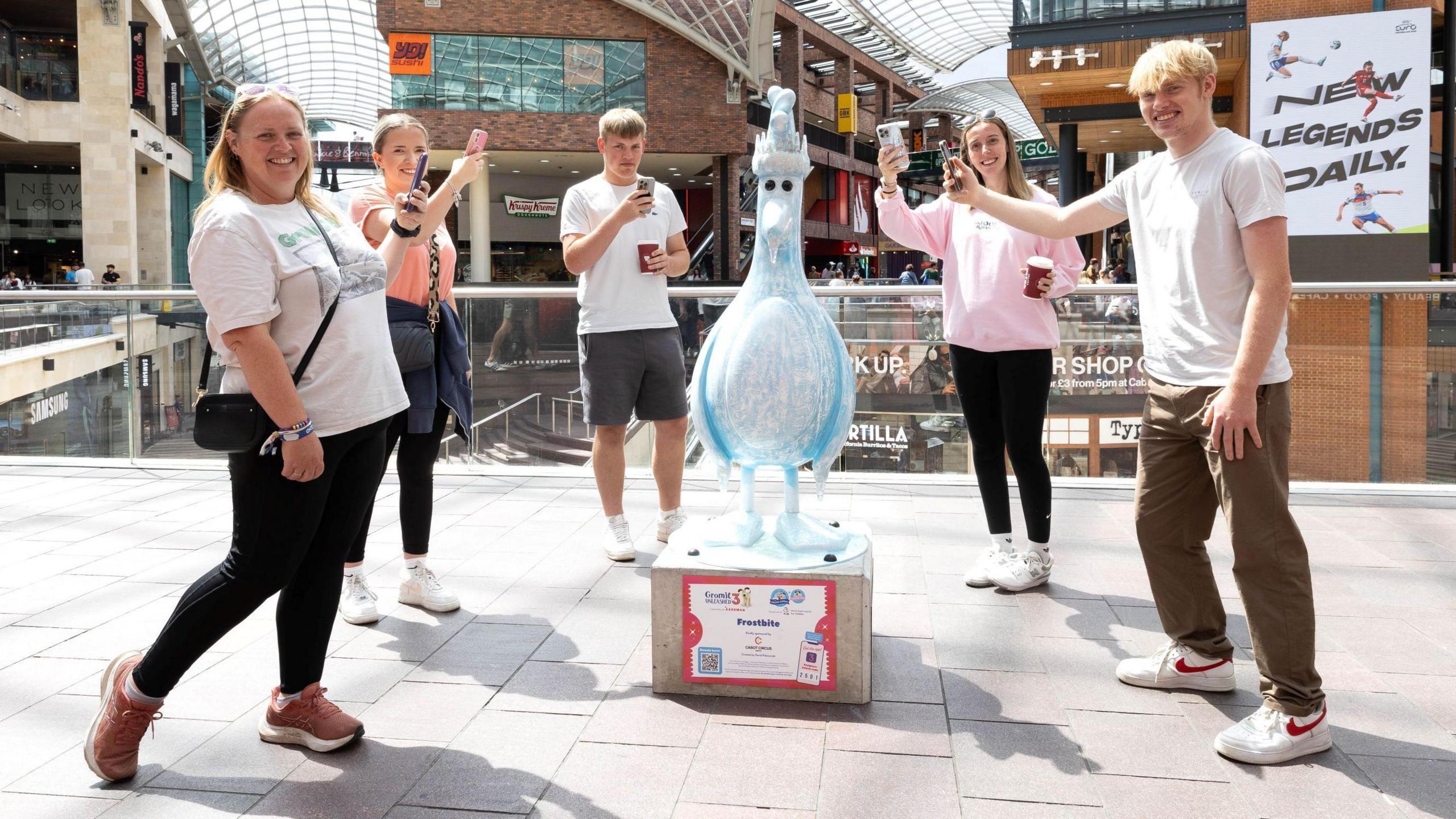 A group of five young people stand around a glass-style statue of Aardman character Feathers McGraw. All the young people are holding their phones out towards the statue, which is inside the Cabot Circus Shopping Centre. Behind them the main heart of the centre is visible.