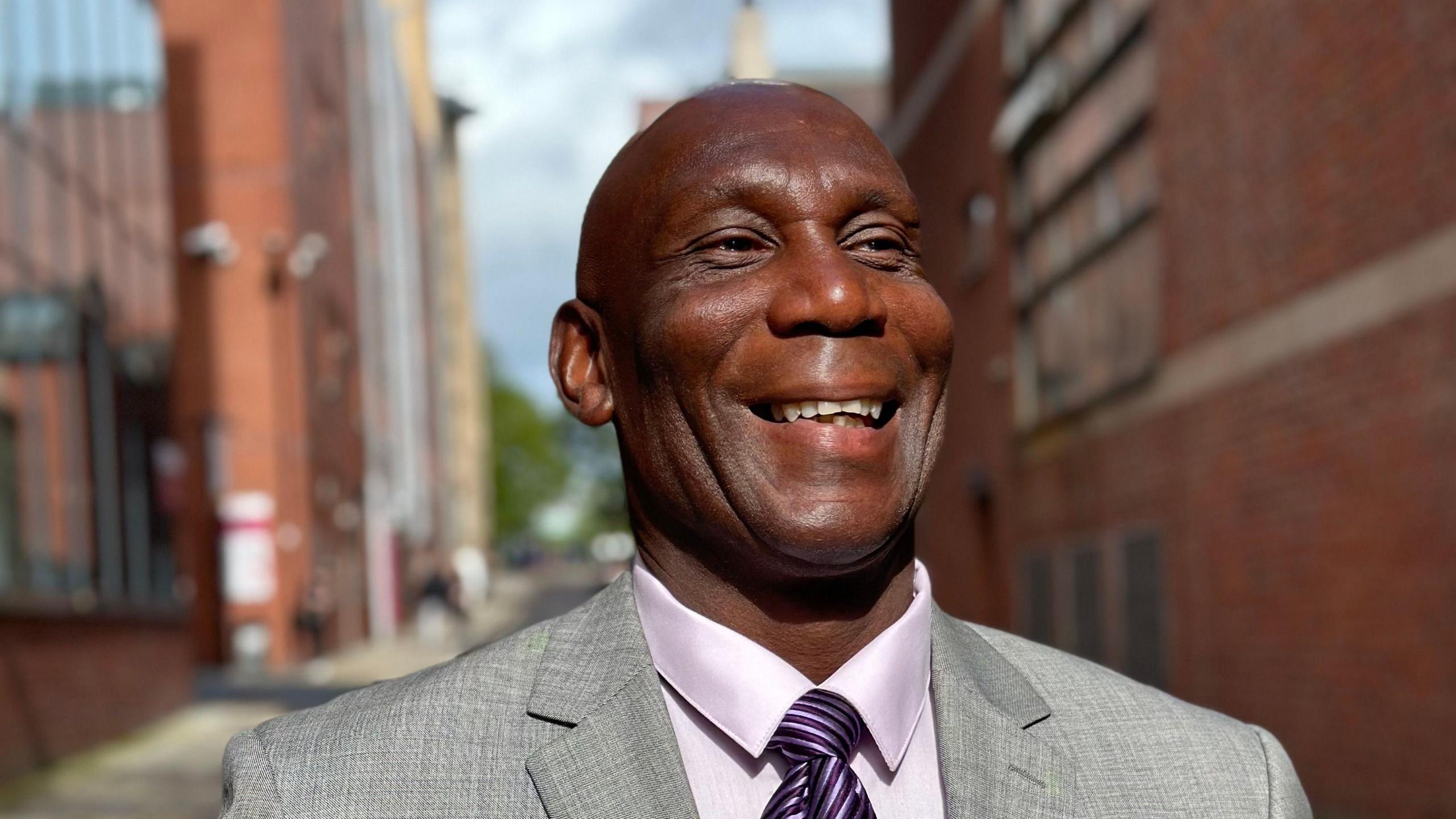 Uriah Rennie, a man wearing a grey jacket, lilac shirt and purple tie. He is smiling and appears to be walking down a narrow street between two large redbrick buildings.