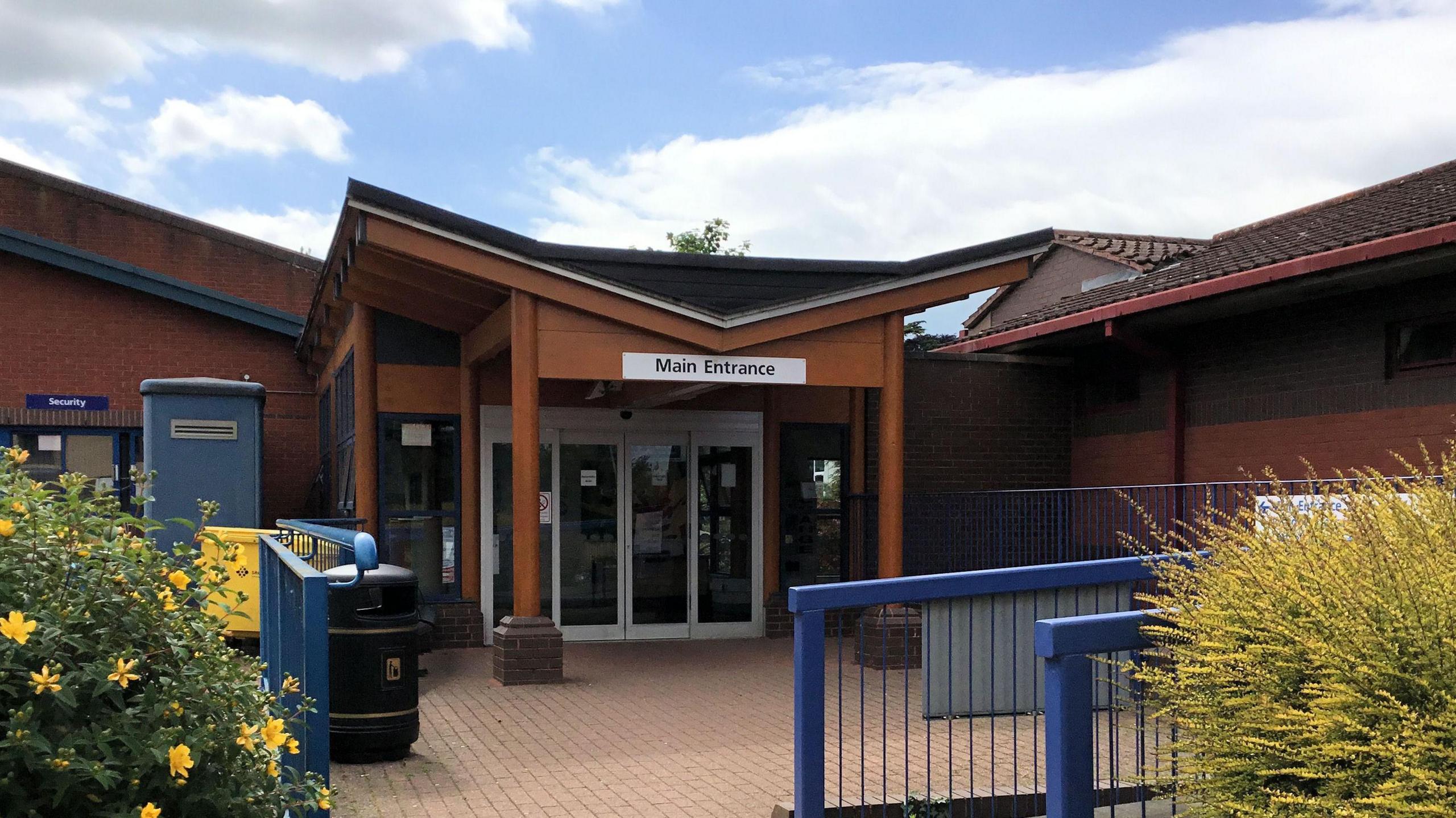 The front entrance of the Horton Hospital. It has a brown wooden canopy over a set of large glass doors. The doors are in the middle of a large red brick building.