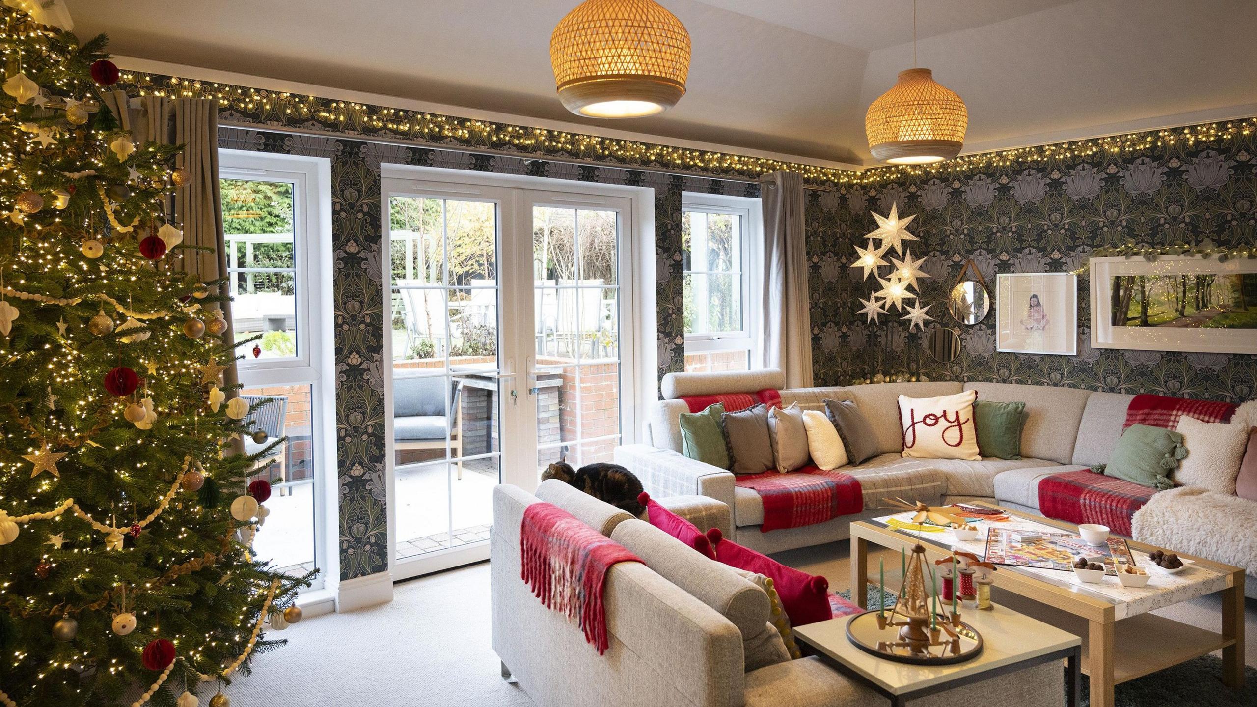 A cosy living room, bordered by fairy lights. A large sofa is covered in cushions, one of which is adorned with the word 'joy'. A large Christmas tree stands in the corner, sprinkled in Christmas baubles and lights. 