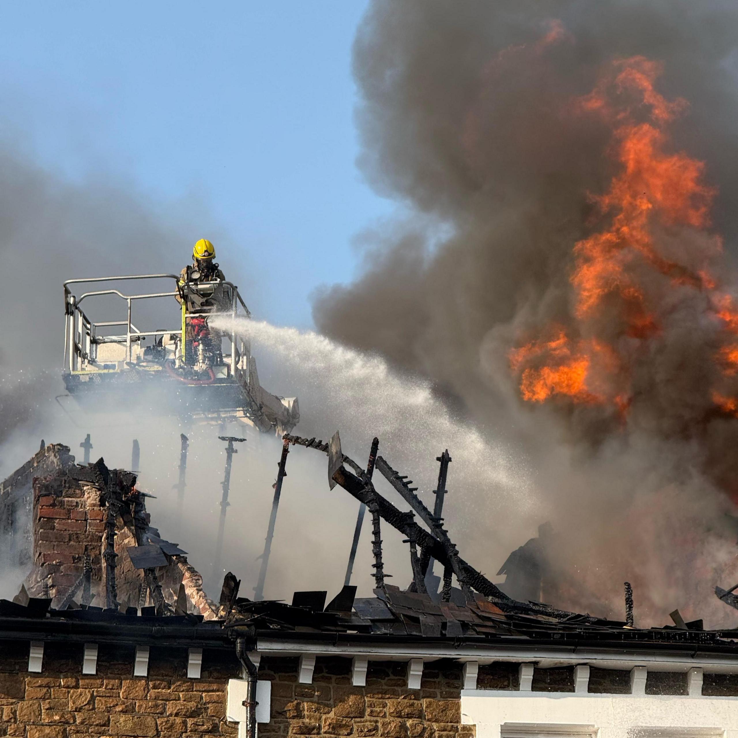 Charred roof of a building with a firefighter spraying water from a platform. Flames and a large amount of smoke can be seen.