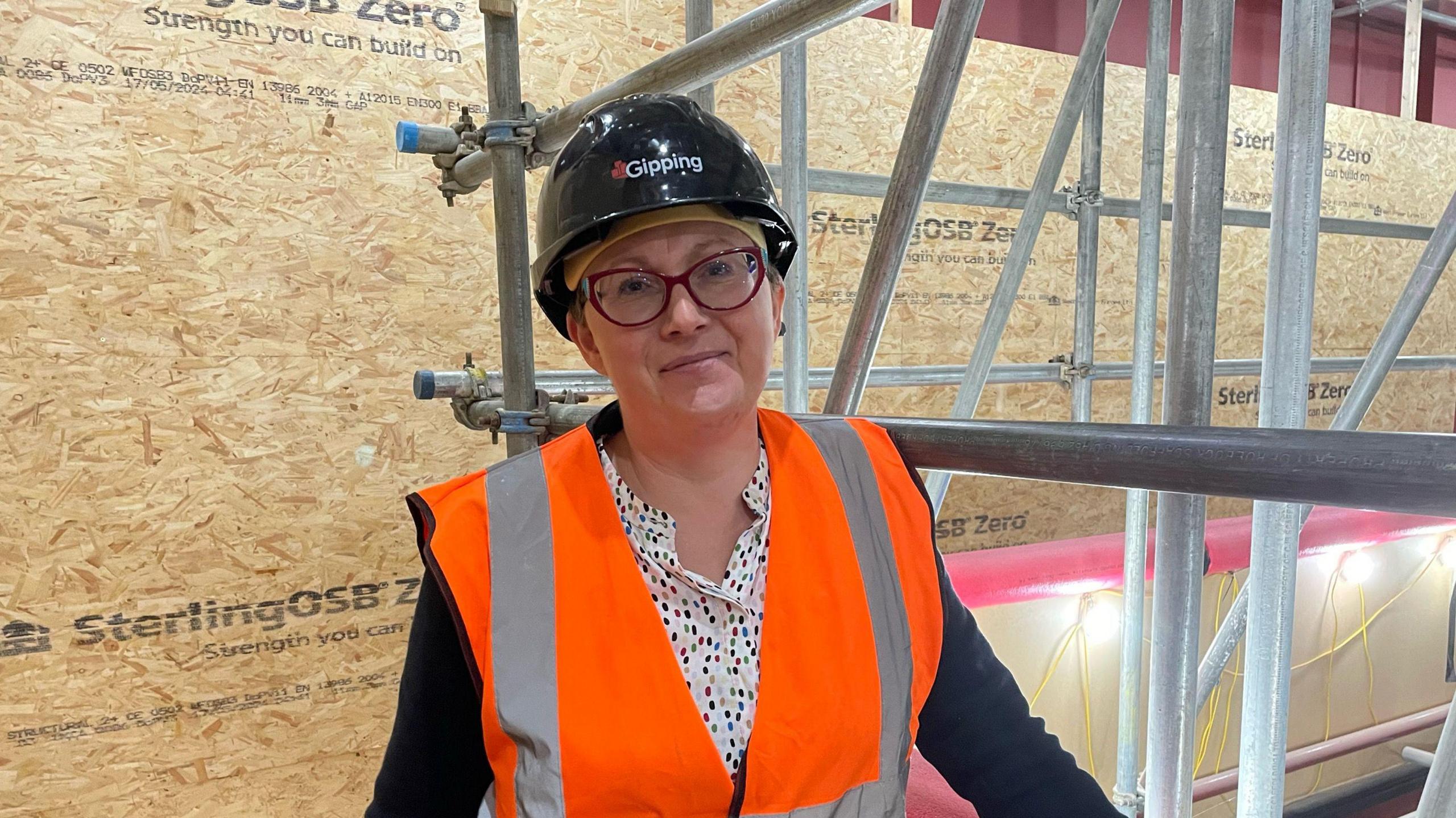 A photo of museum manager Alison Hall. She is smiling to camera while wearing a black safety helmet and orange high-vis jacket. Around her is scaffolding along with plywood construction boards protecting the museum's walls.