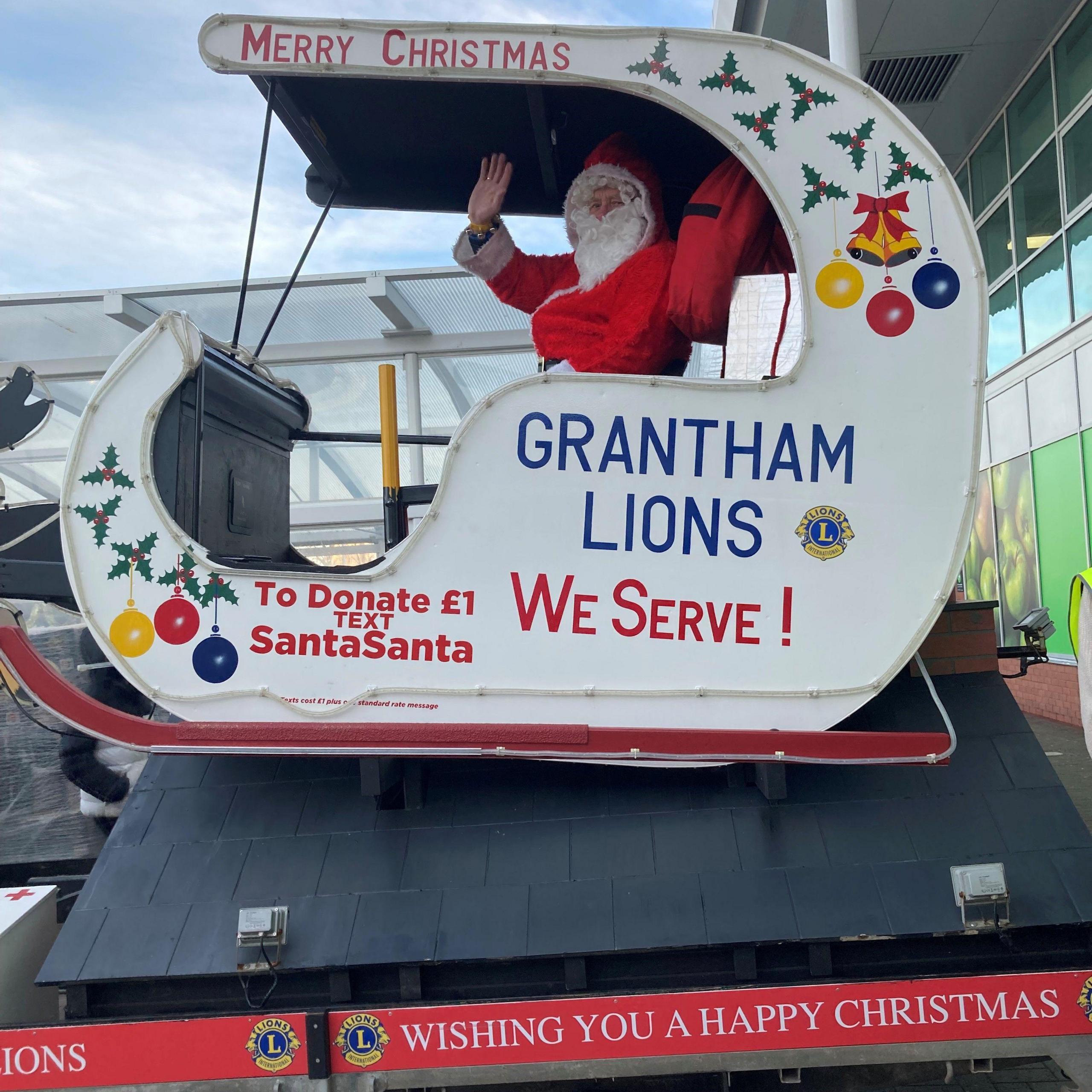 White Santa sleigh during the day. Santa is waving from the sleigh. On the side of it it says "Grantham Lions. We Serve! To donate text SantaSanta." It also says "Wishing you a happy Christmas".