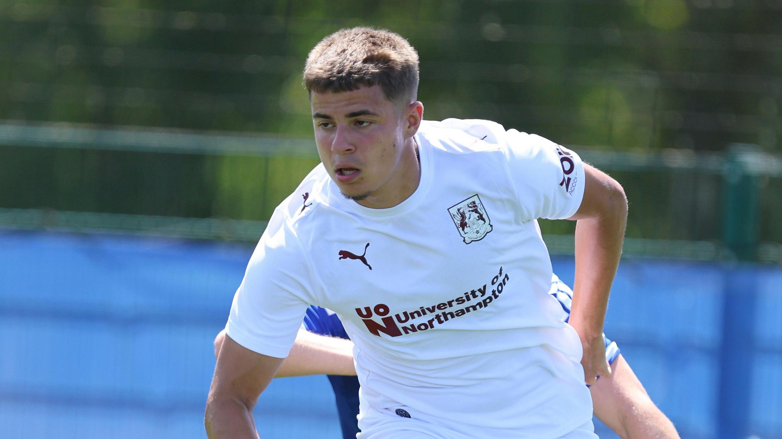 A young 20-year old footballer, wearing a white football shirt with the University of Northampton logo printed on the front