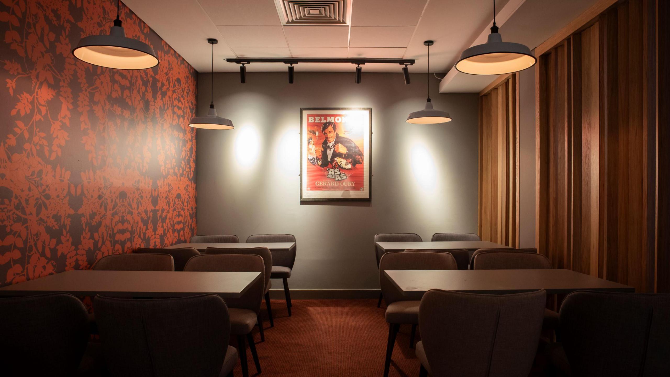 A stylish room with tables and chairs on either side, a moody orange and brown colour scheme and a large movie poster on the far wall.