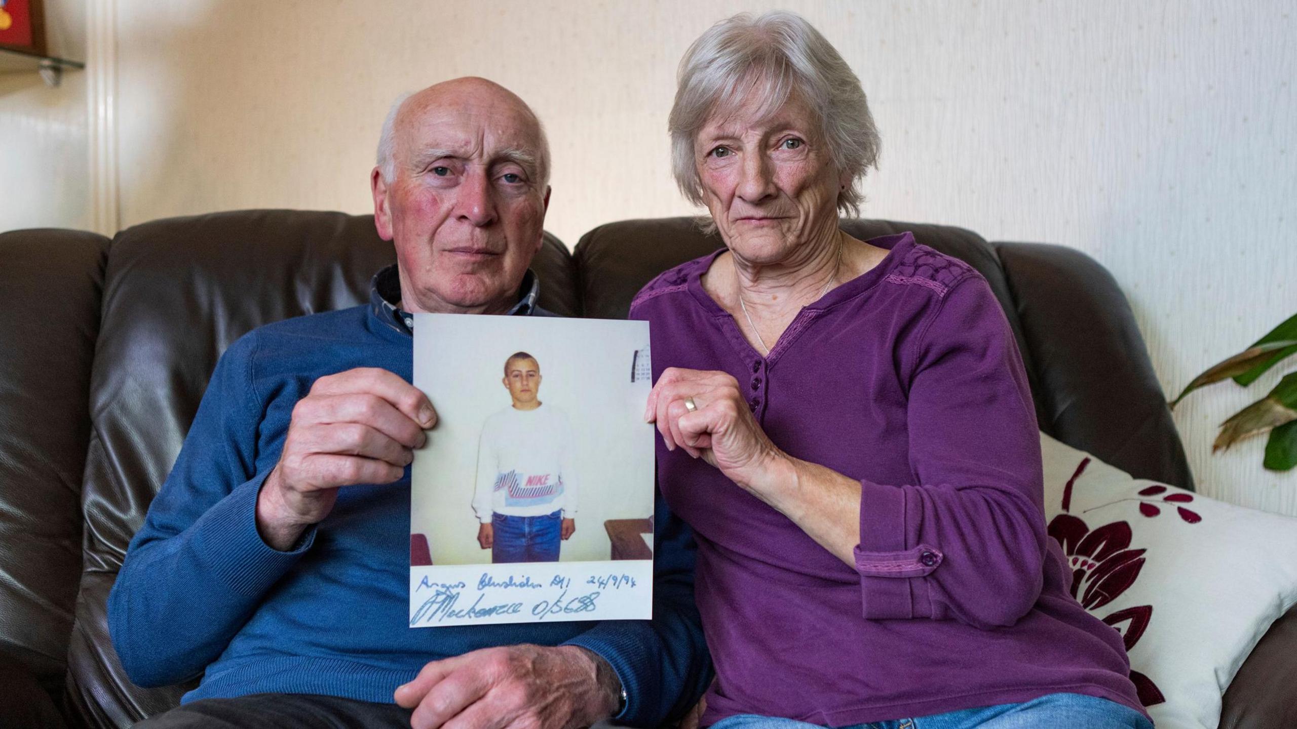 Michael Ross's parents sitting on a brown, leather sofa holding up a photograph of him when he was 16. He has a bald head and is wearing a blue jumper over a shirt and she is wearing a purple top and jeans