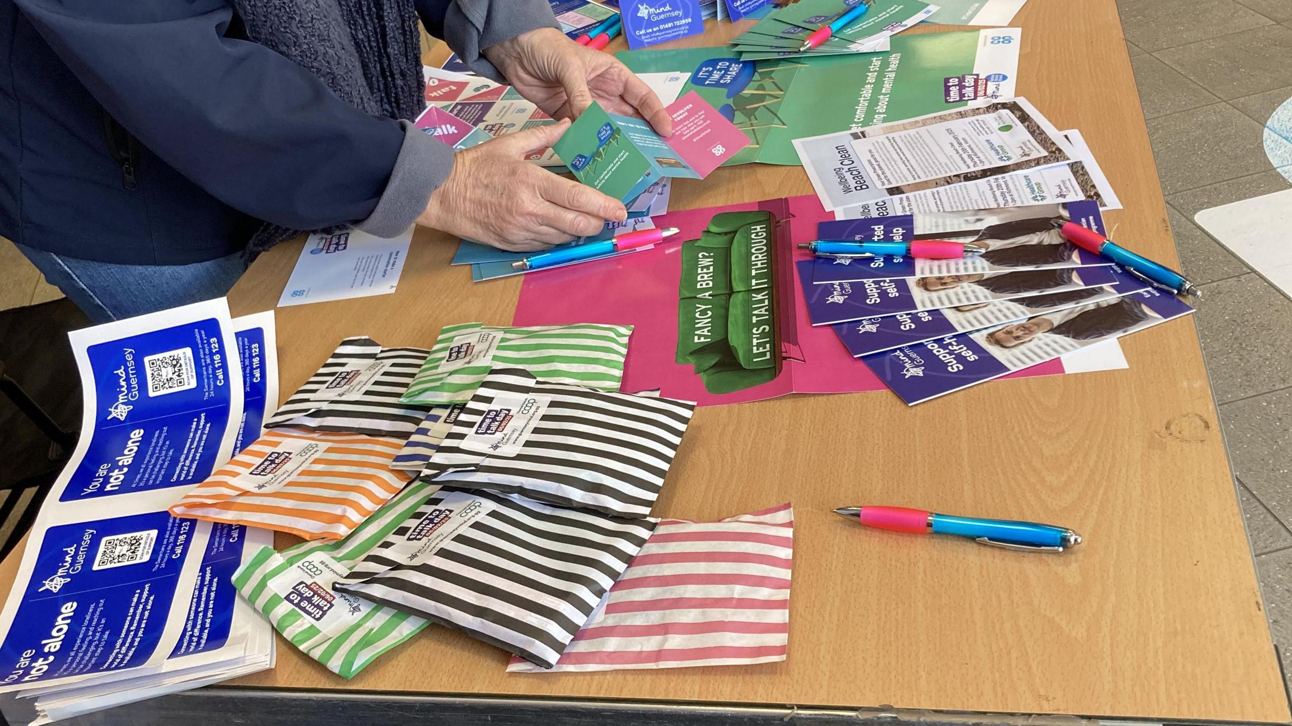 A pair of hands holds a multicoloured pamphlet, about a table covered in goodie bags, leaflets, and a pen.