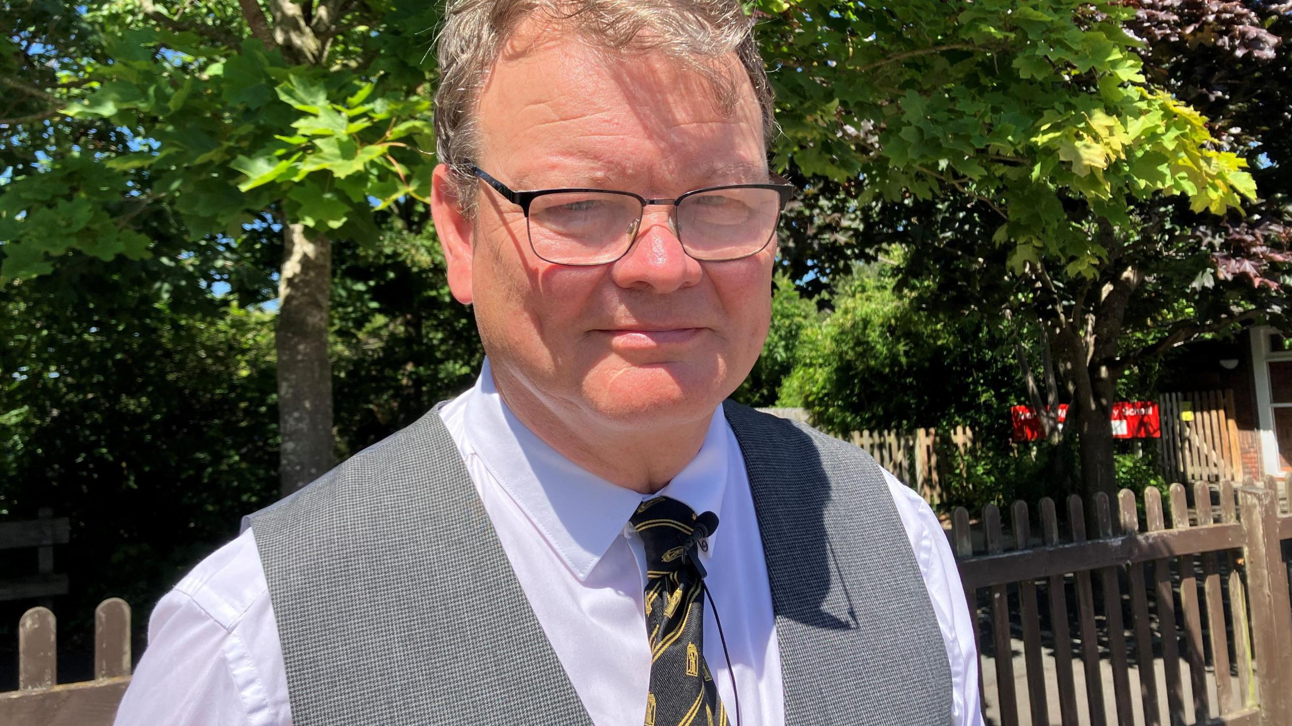 A man with glasses and grey hair looks to the camera and smiles. He is wearing a white shirt, grey waistcoat and dark tie with yellow stripes. He is beside a fence and tree and is in the sunshine.