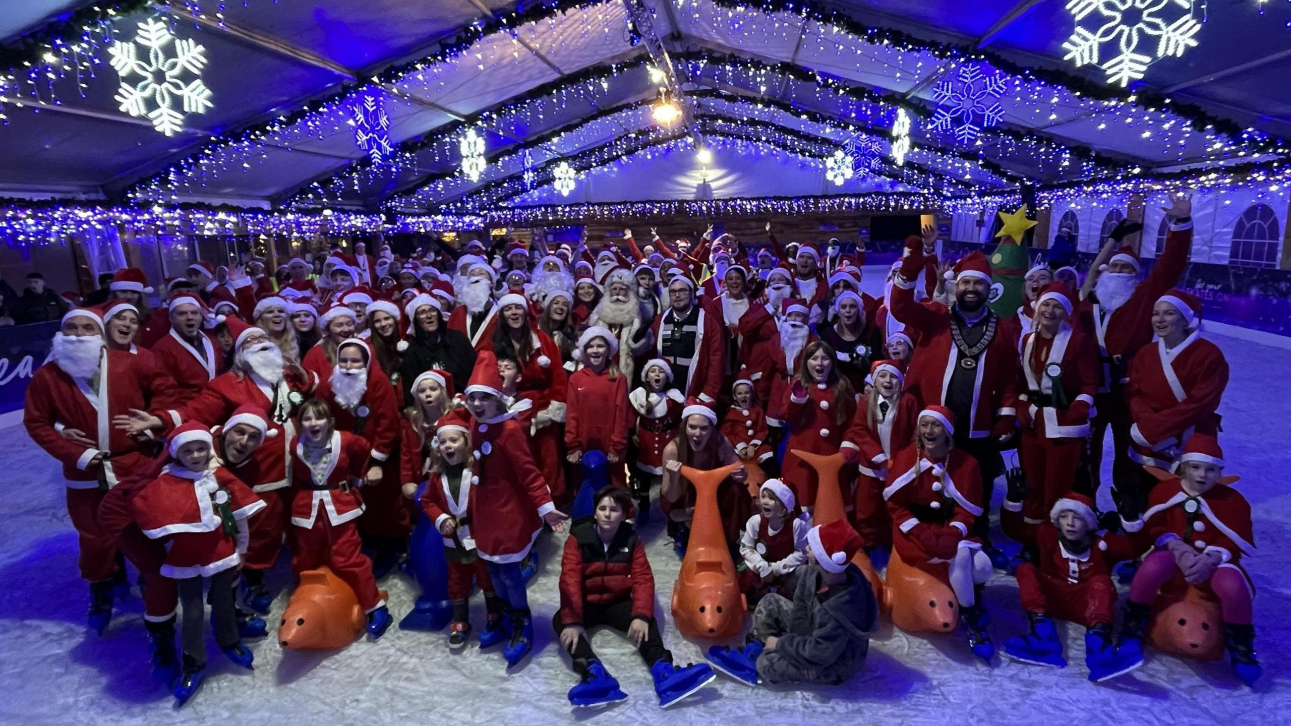 A group photo of people dressed as Santa on an ice rink at Clarks Village in Street. Some of them are seated on the ice and many are standing and smiling at the camera. The picture is taken at night and the rink is lit up by illuminations overhead, some of them shaped like snowflakes