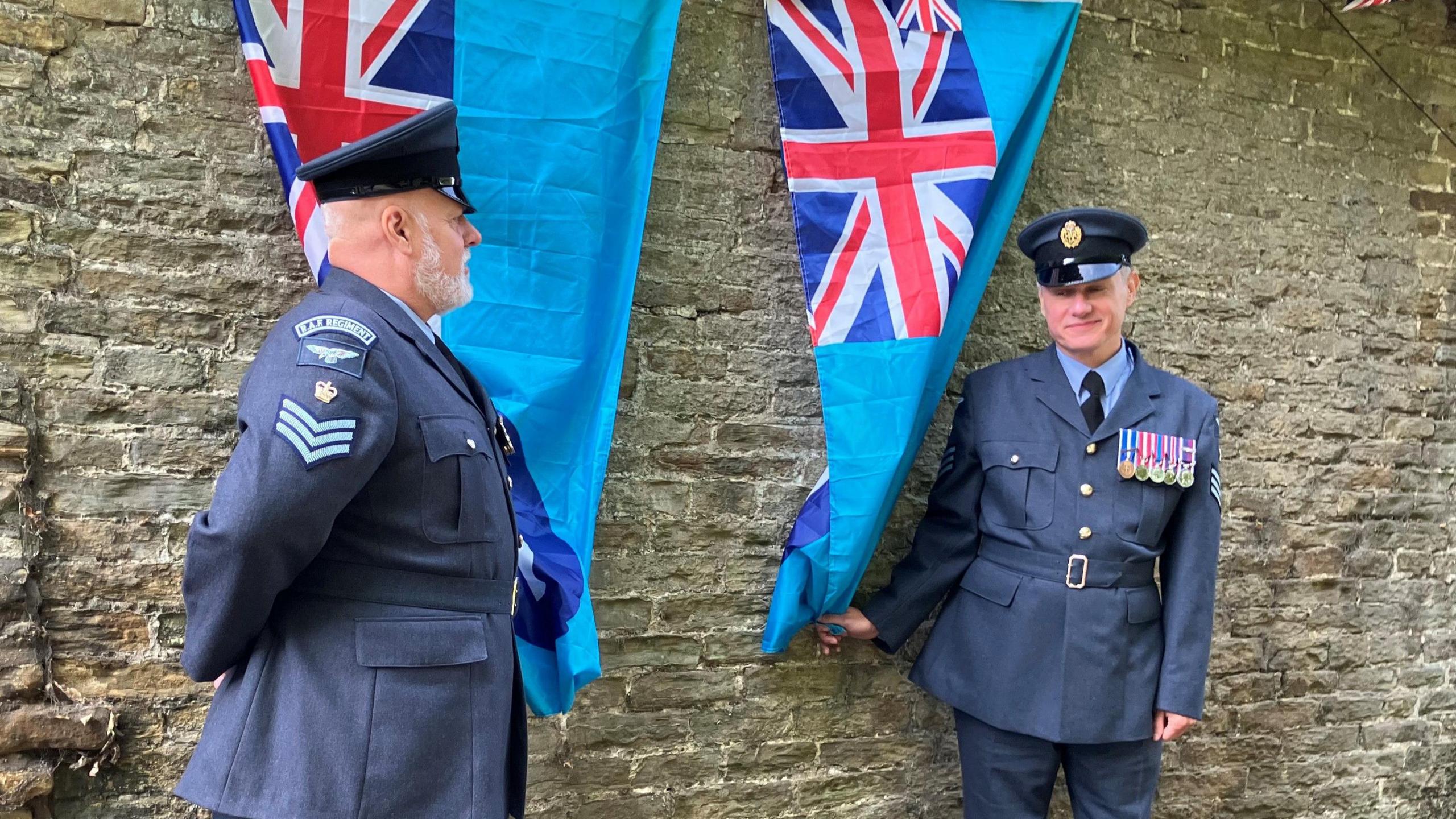 Two men in RAF uniform stand in front of a brick wall. A light blue flag, with a target on one side and the Union flag in the corner, hangs behind each of them. Each man holds a flag, ready to pull it aside to show the plaques underneath.