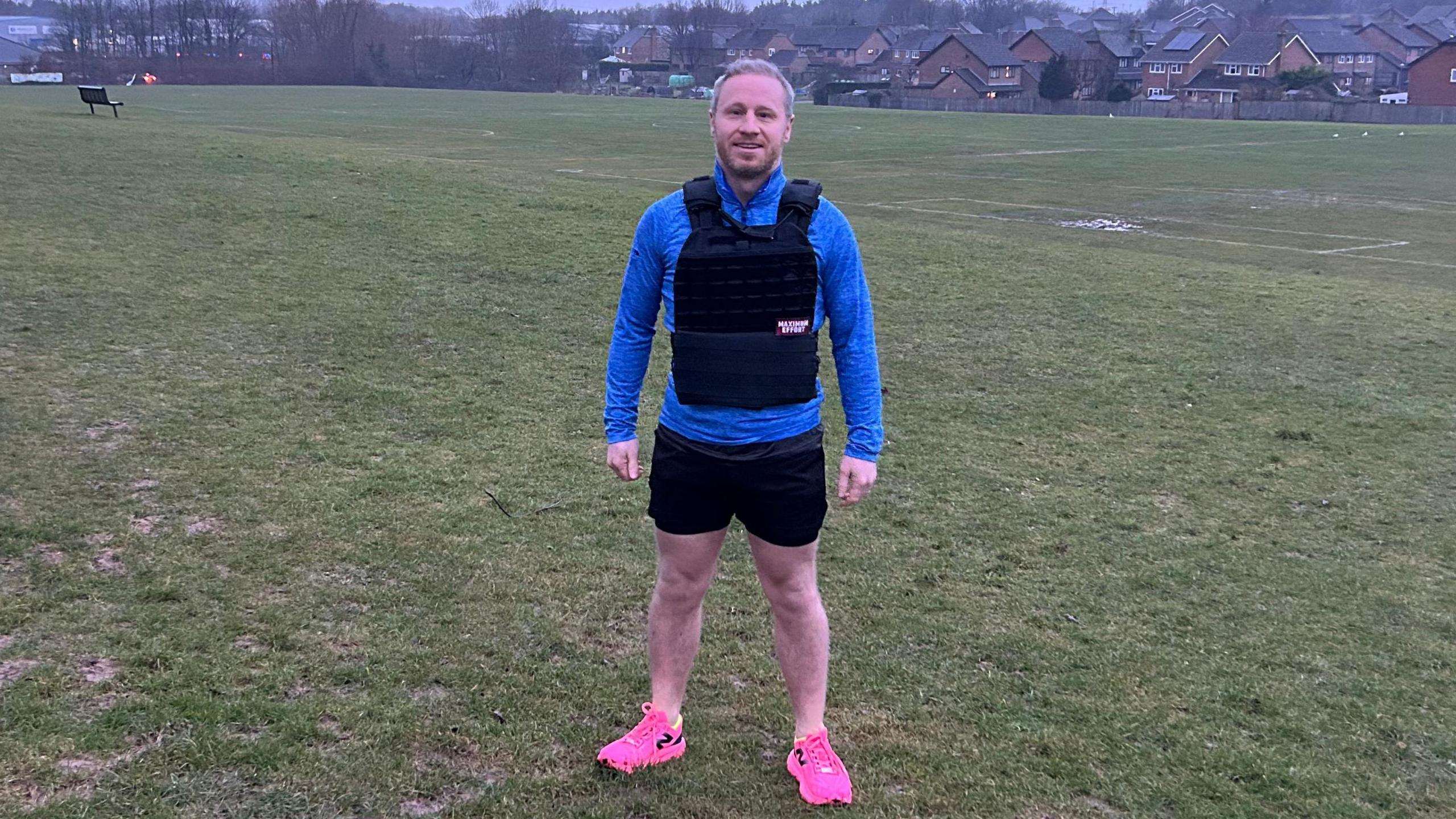 Pete Grimwade in his marathon running gear of blue, longsleeved top, black shorts and pink trainers, wearing his 10kg pack strapped to his chest - standing in a field 