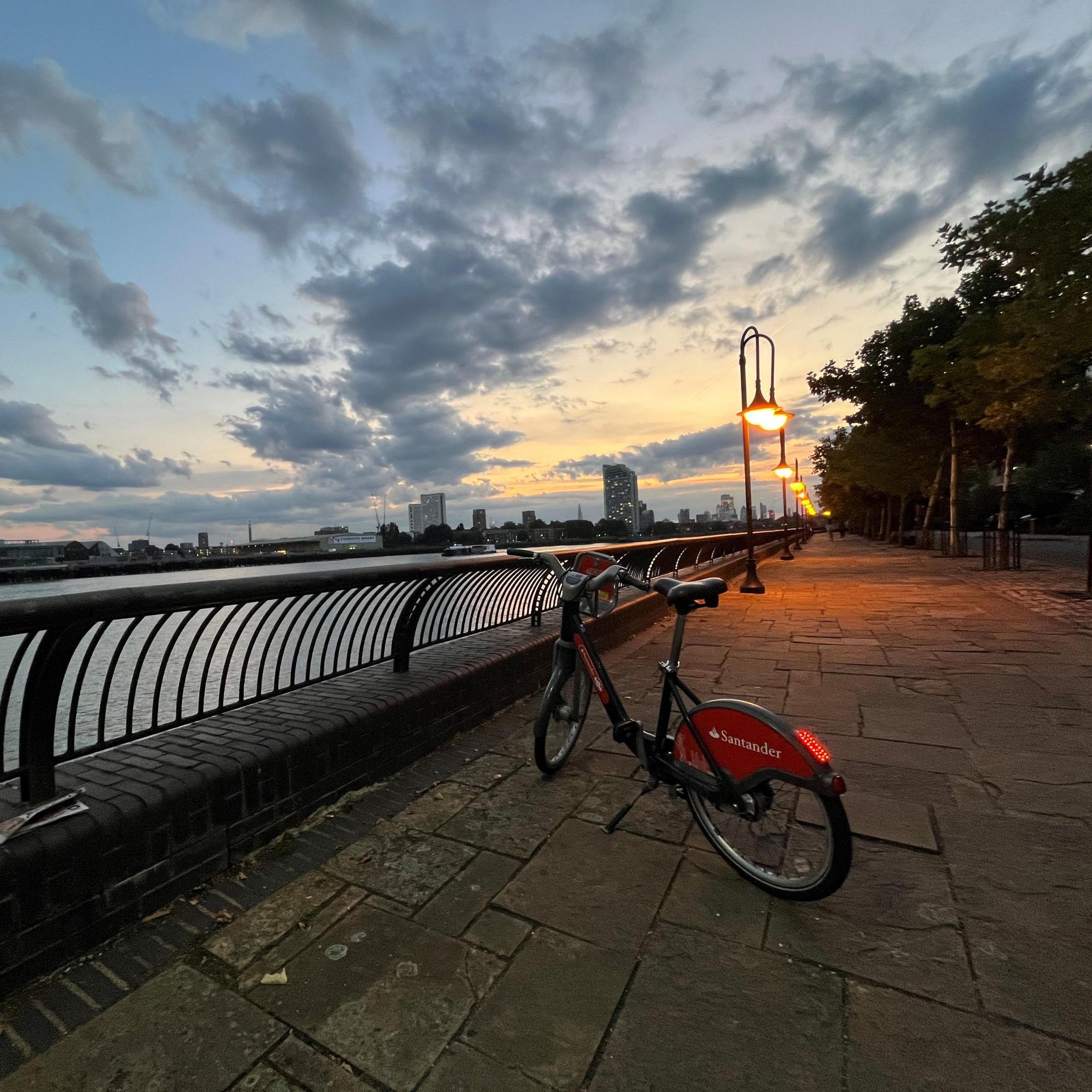 A Santander Cycle stands on a stone riverside path beside black railings. Streetlamps line the walkway and glow orange as the sun sets. The Thames is in the background with tower blocks and cranes on the opposite bank.