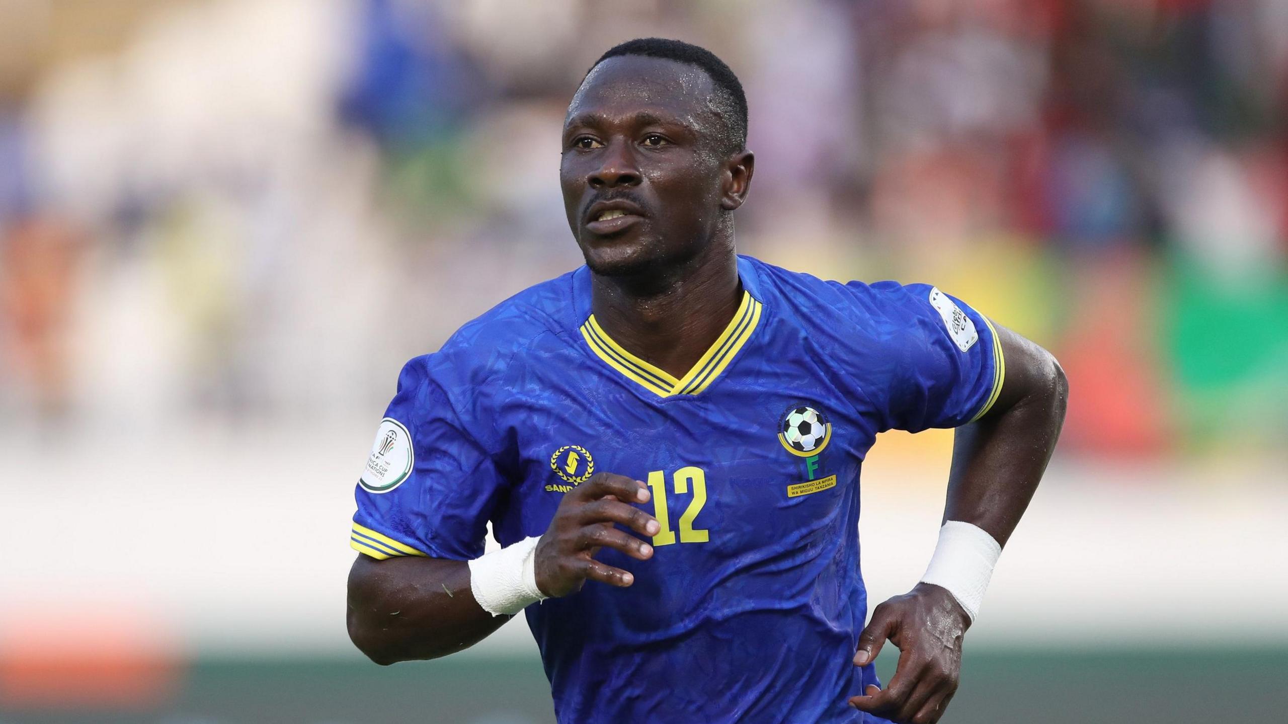 Simon Msuva, wearing a blue Tanzania shirt with yellow trim and the number 12 on the front, runs towards the camera during a football game