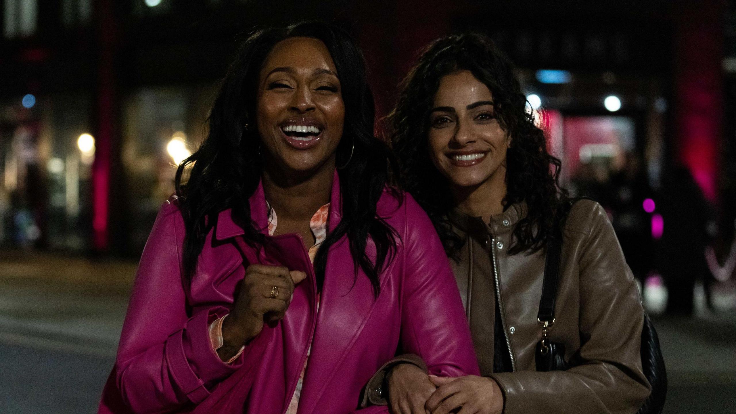 Alexandra Burke is wearing a magenta pink jacket and is stood next to Mandip Gill wearing a brown jacket. It is night-time and the two are smiling and laughing at the camera.