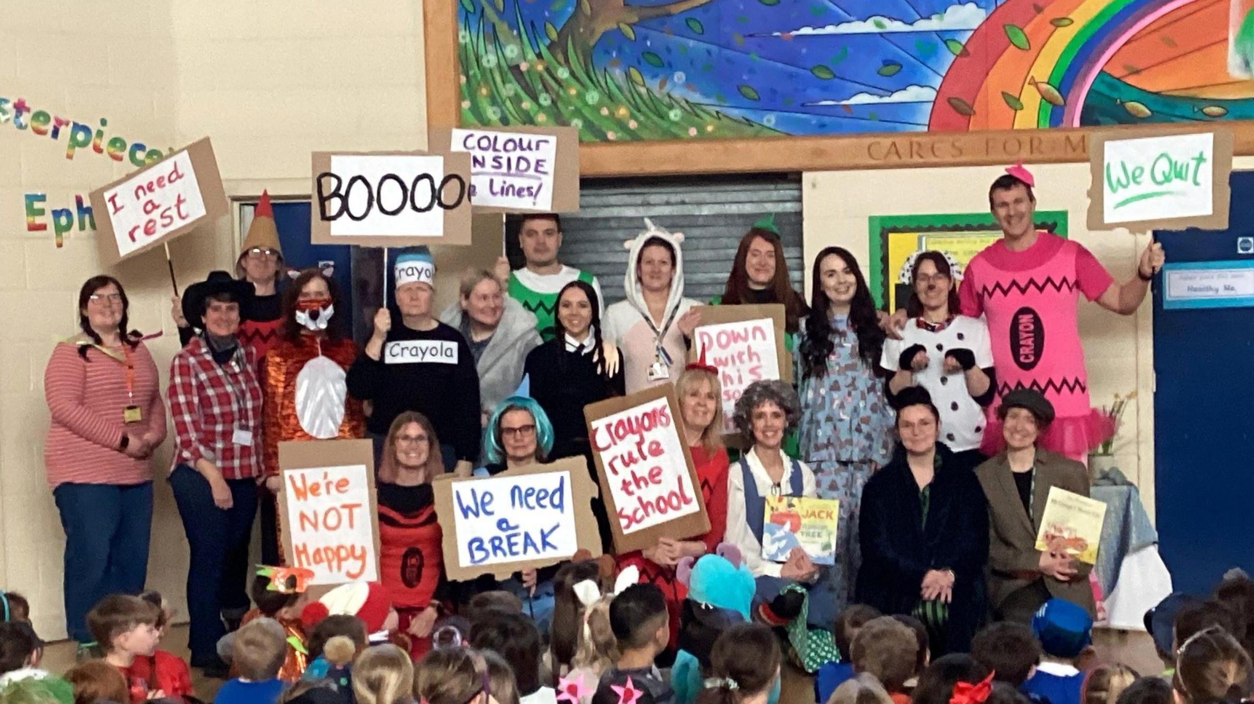 A large group of staff at St John's School in Clevedon stand facing pupils during an assembly on World Book Day. The staff are in different types of fancy dress and holding up placards saying they are fed up with work and are going on strike