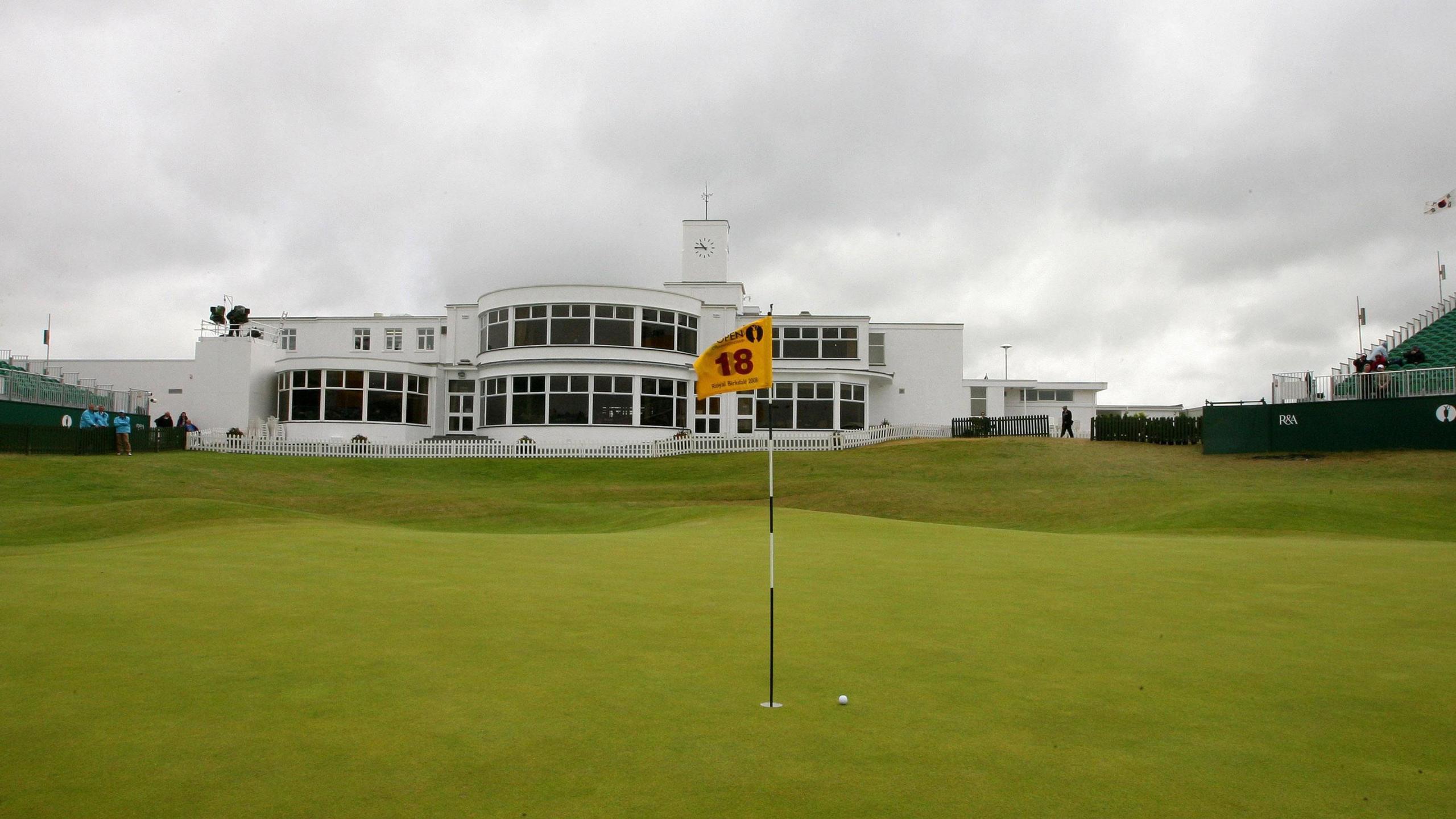 A golf course, in the foreground is the green, flag and a golf ball near the hole. In the background is a large white building. On the edges of the image are the start of stands on either side of the course.